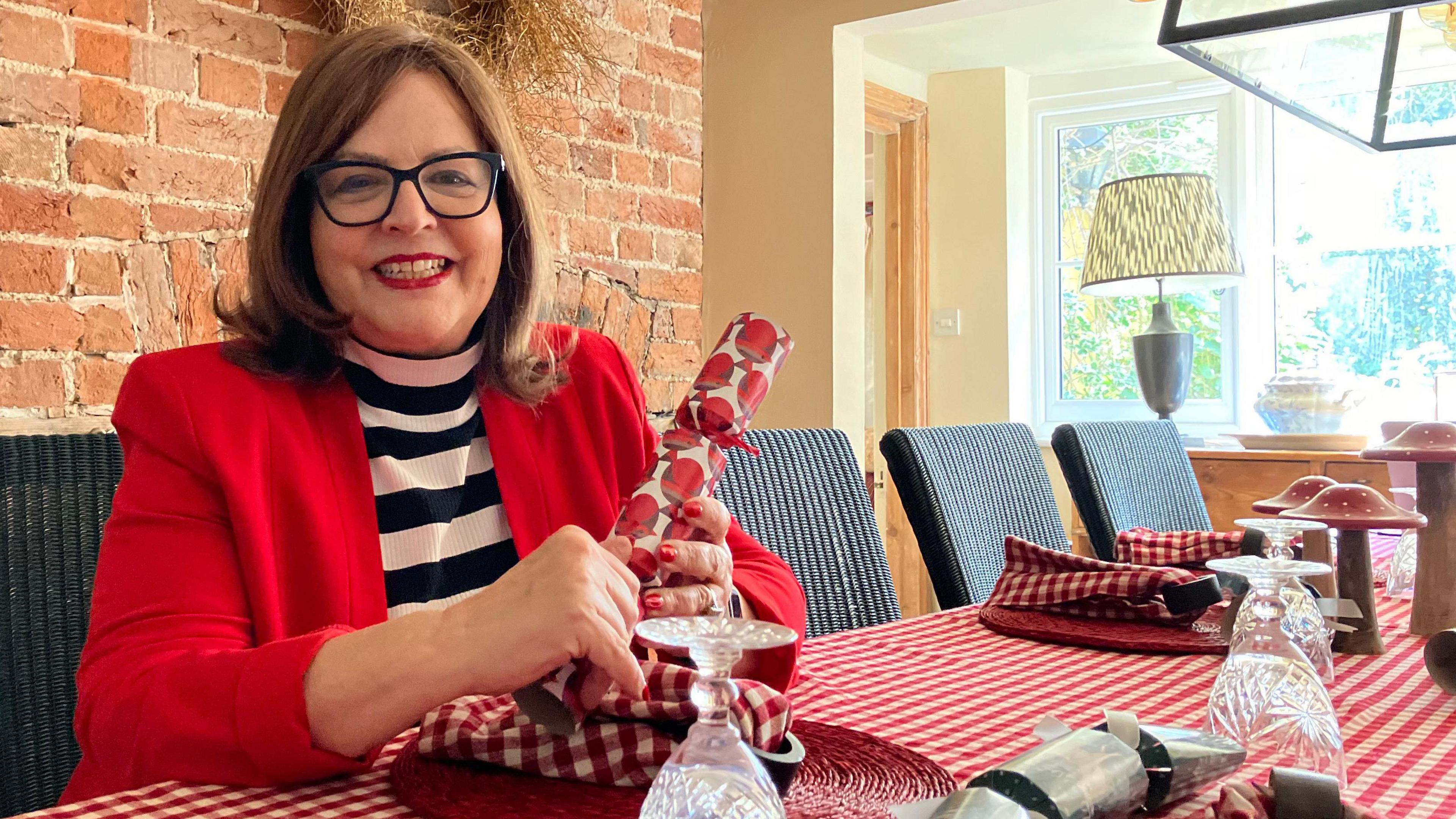 Jane Malyon sat at the table holding a cracker. The table cloth has red and white checkers and has napkins, crackers and glasses on it. Mrs Malyon herself is smiling at the camera.