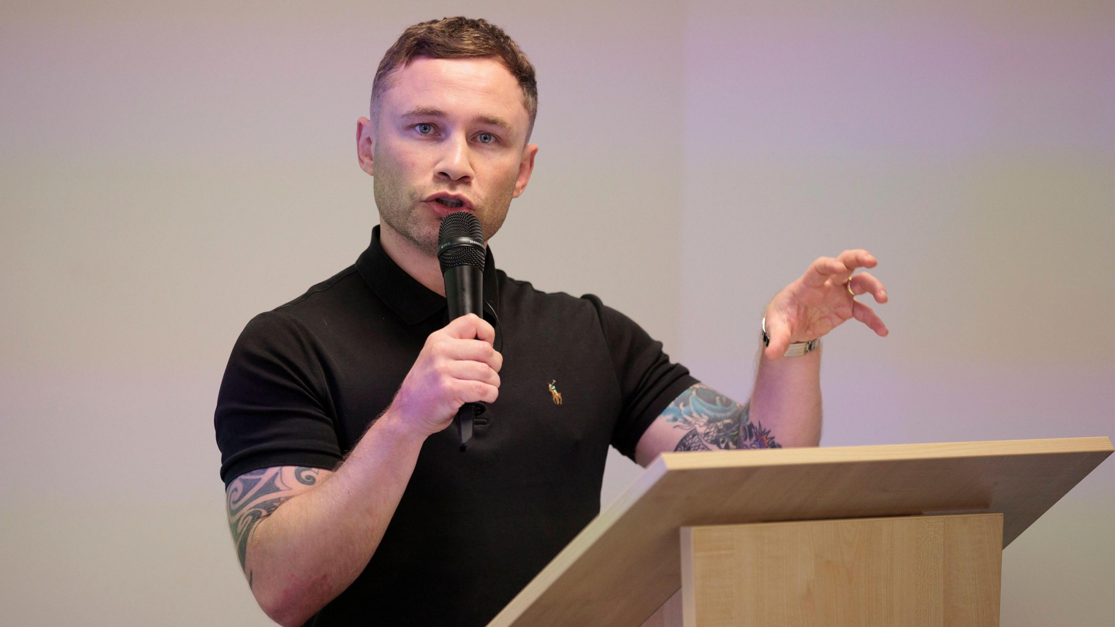 A man dressed in a short sleeved black top stands at a wooden podium