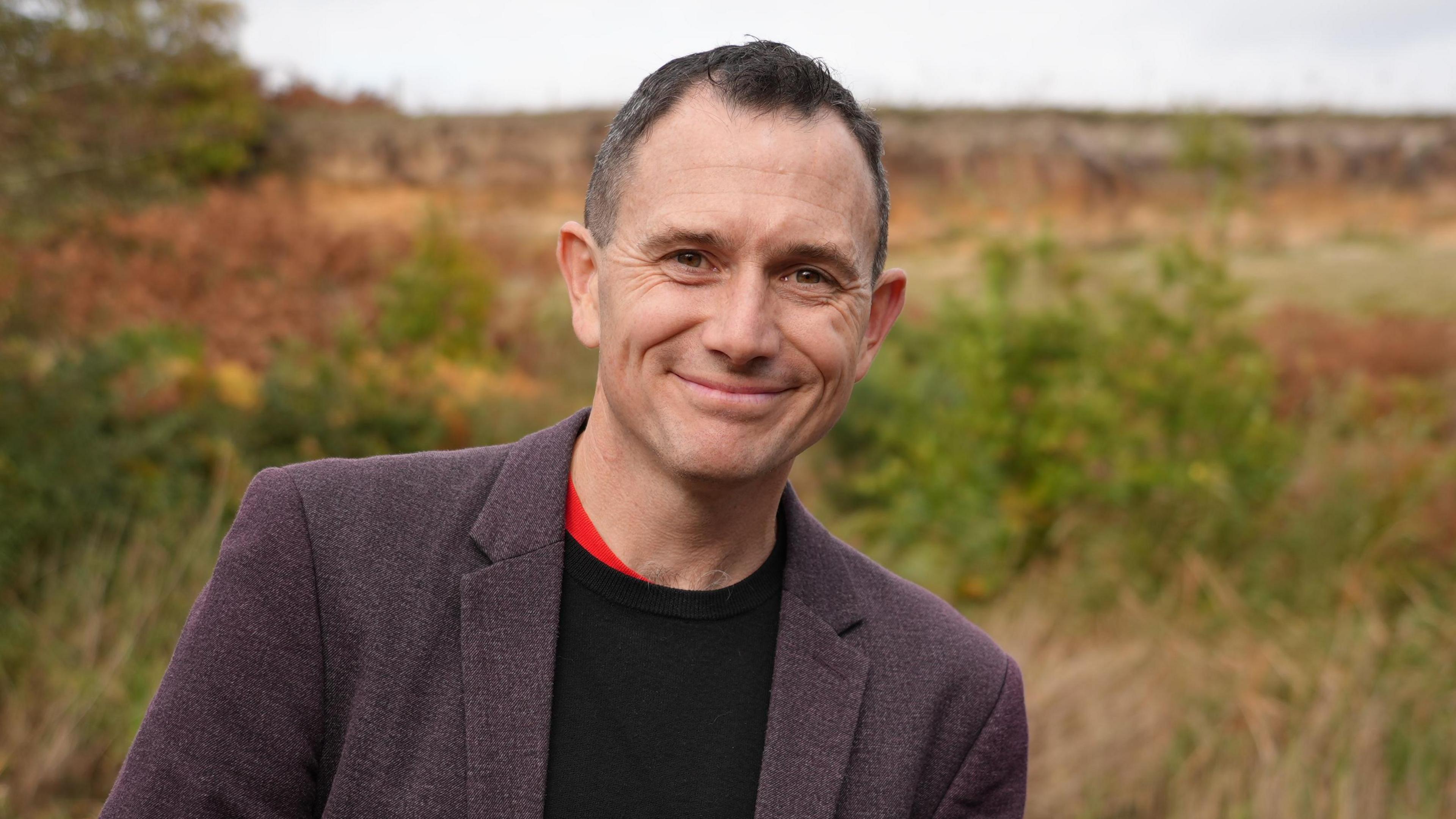 Tom Hall is wearing a charcoal coloured blazer with a black shirt underneath. He has short dark hair and is smiling warmly at the camera with  RSPB Minsmere visible behind him