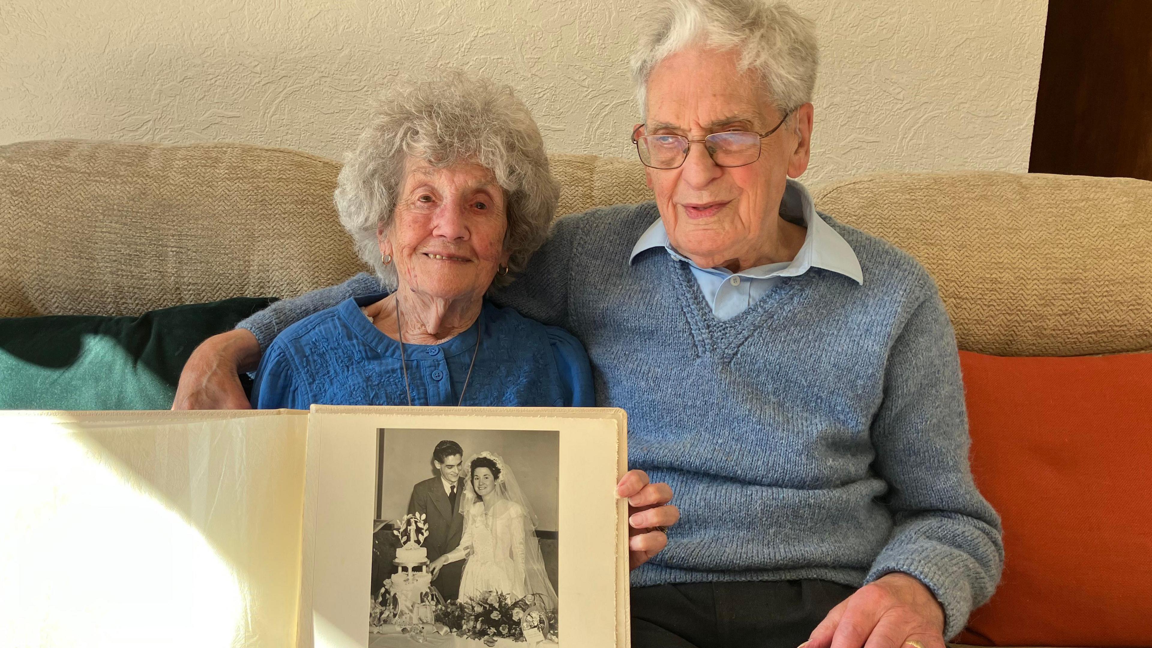 An elderly couple sit on a cream sofa together while the woman in a blue top holds up a photo album showing a black and white photo of them when they were younger cutting their wedding cake. The man is sitting in a grey jumper with his arm around the woman. 