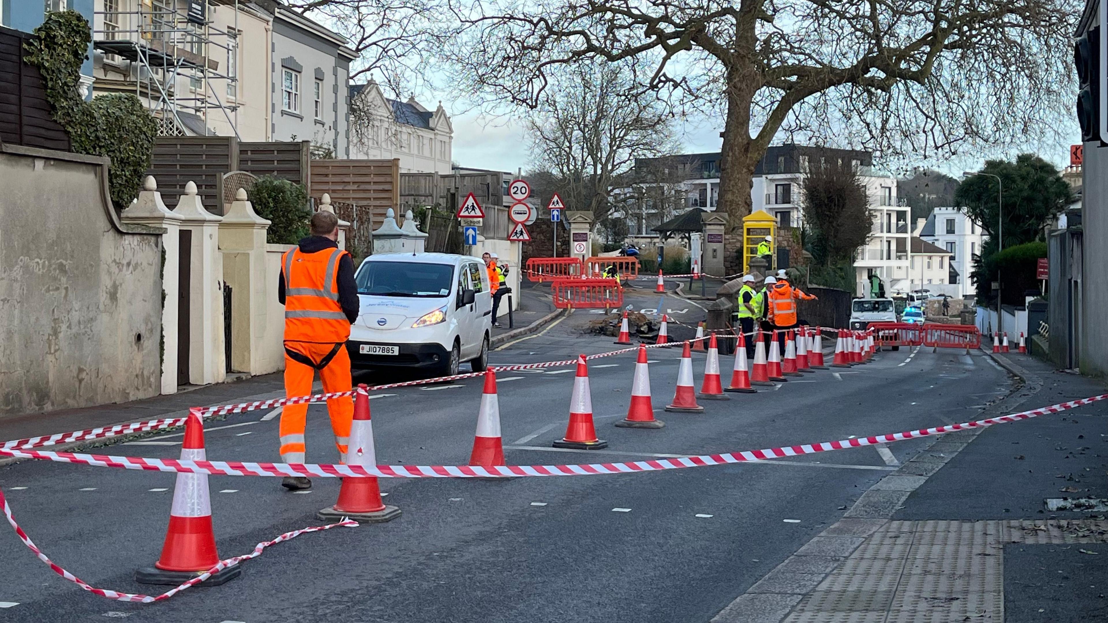 A picture of the scene where the sinkhole is. Cones can be seen separating a section of the road off. There are workers walking around with tape.