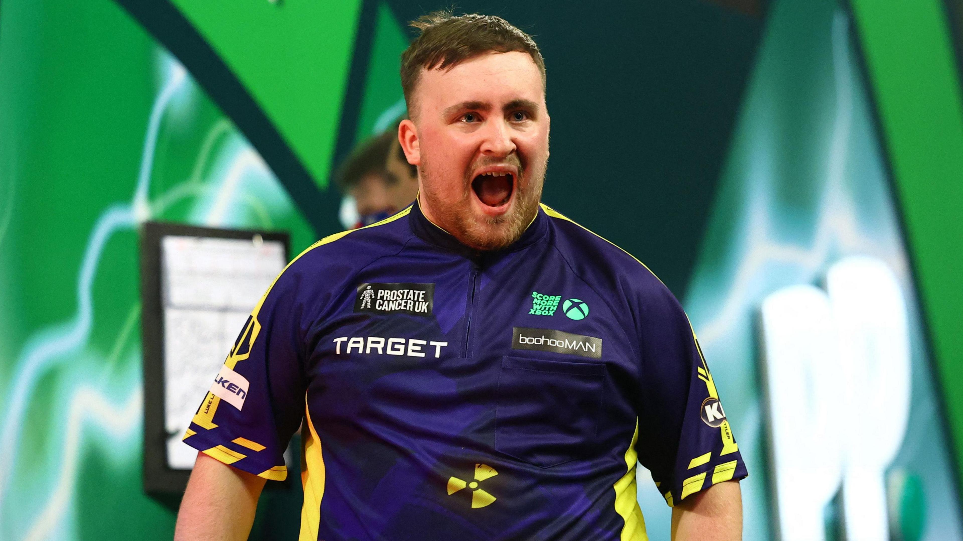 Luke Littler has his mouth open wide in a smile as he looks off from camera on the darting stage 