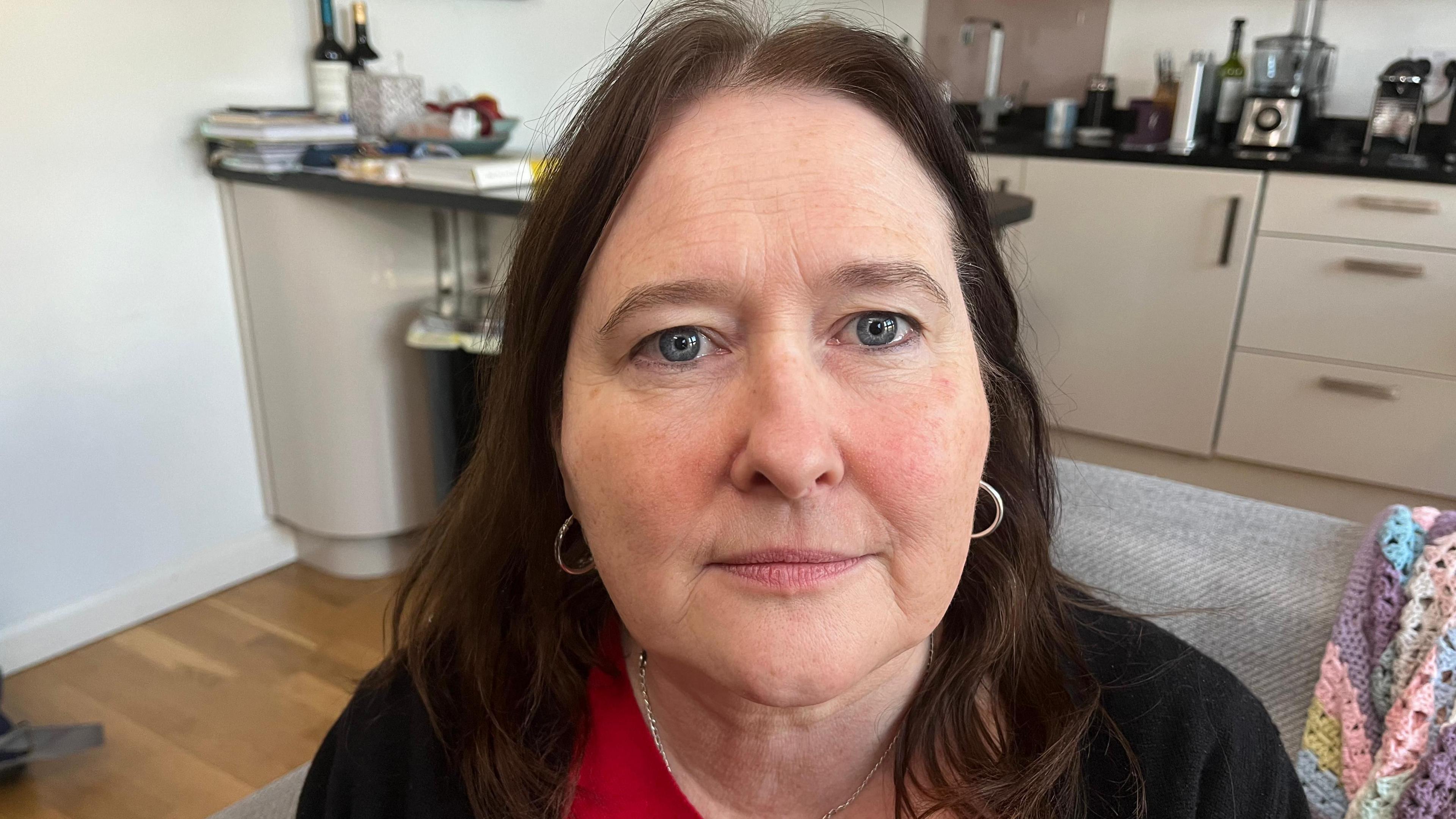 A woman with shoulder-length dark hair wearing a red top and black cardigan sits in a kitchen and living space.