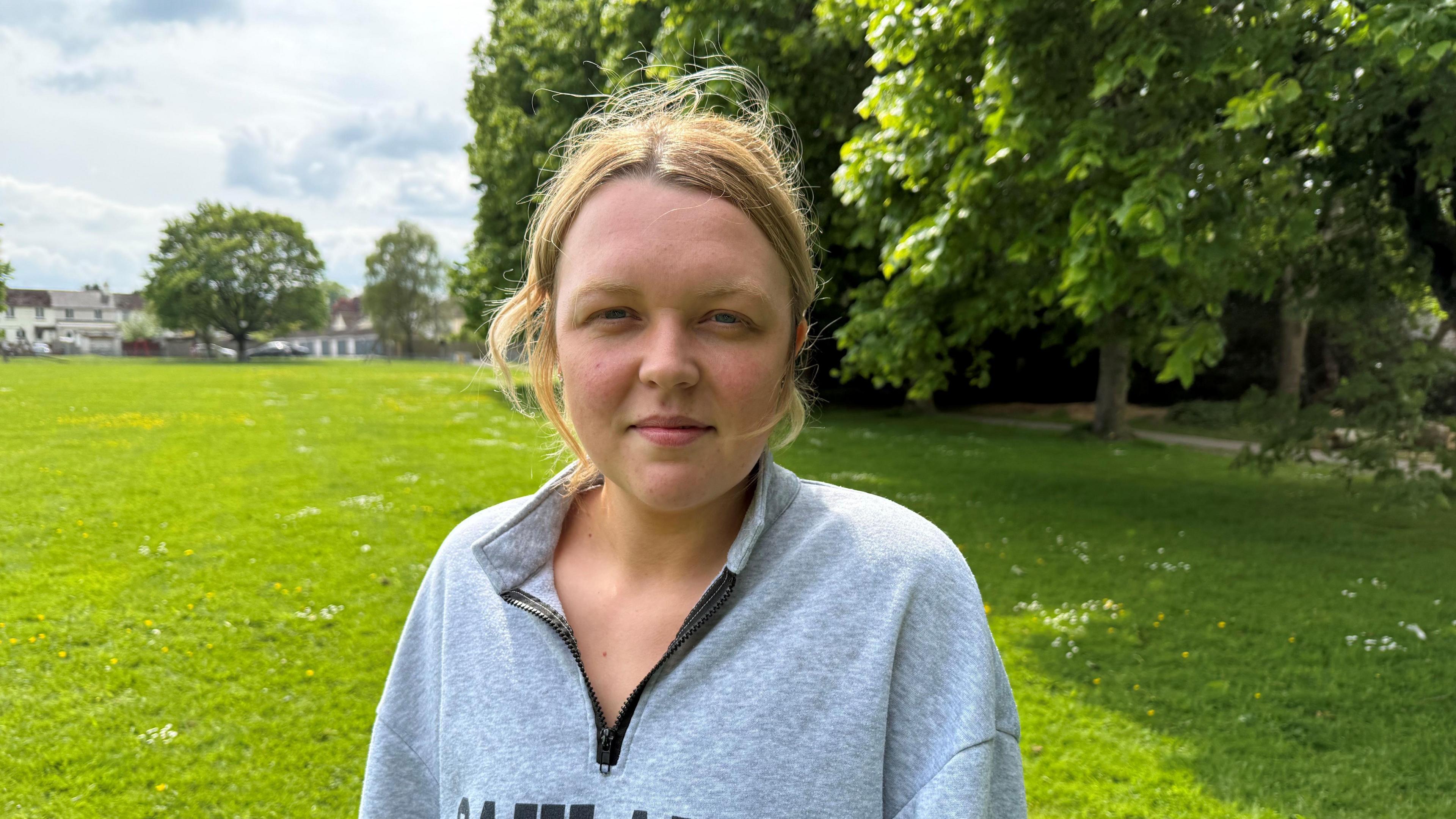 Blonde haired women stood looking at the camera in a green park