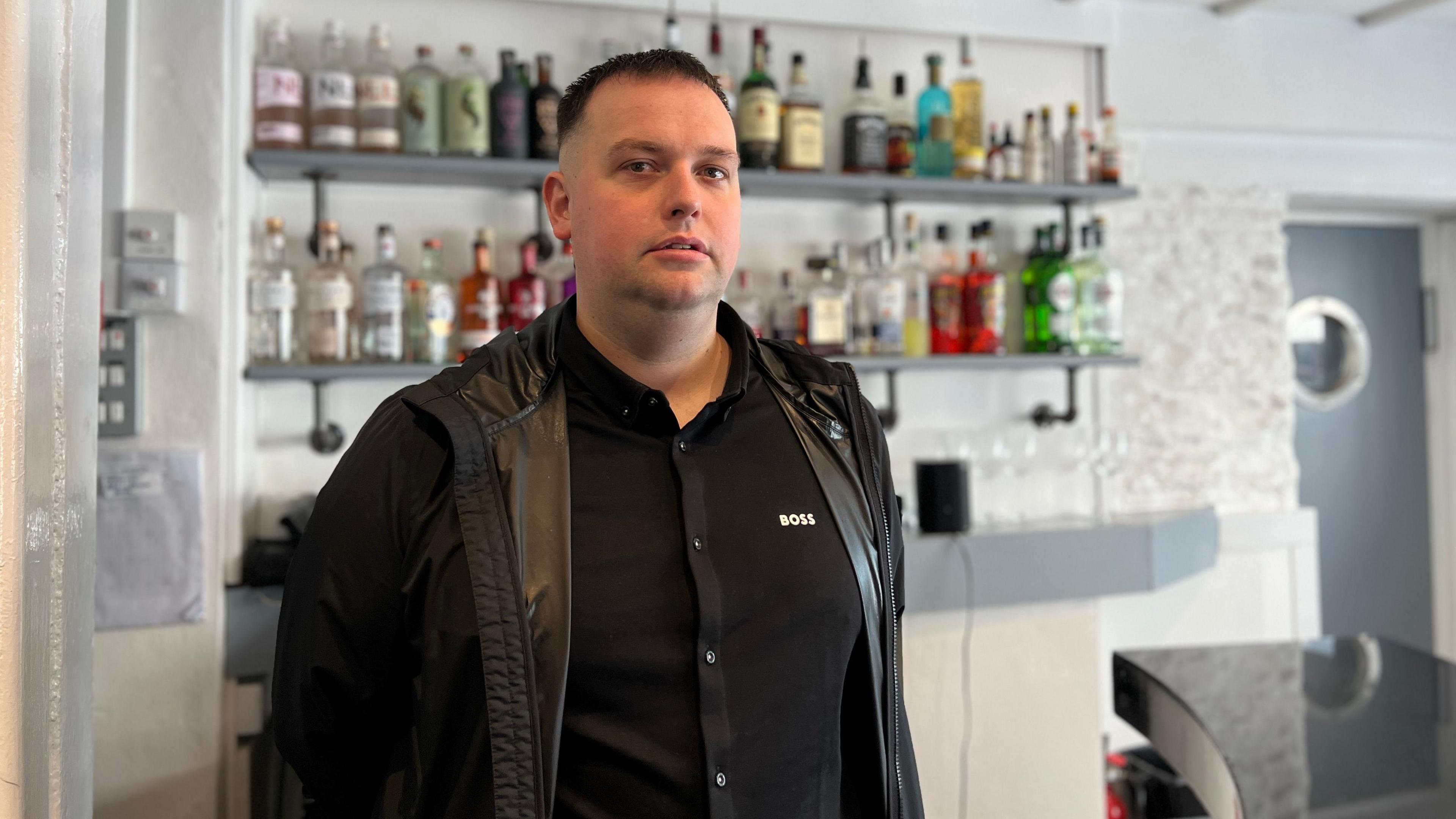 Jamie is looking at the camera and not smiling, standing in front of two shelves full of bottles of spirits wearing a black 'Boss' shirt and black jacket. Part of a black bar top is visivle in the bottom corner and a grey door with a porthole is in the background.