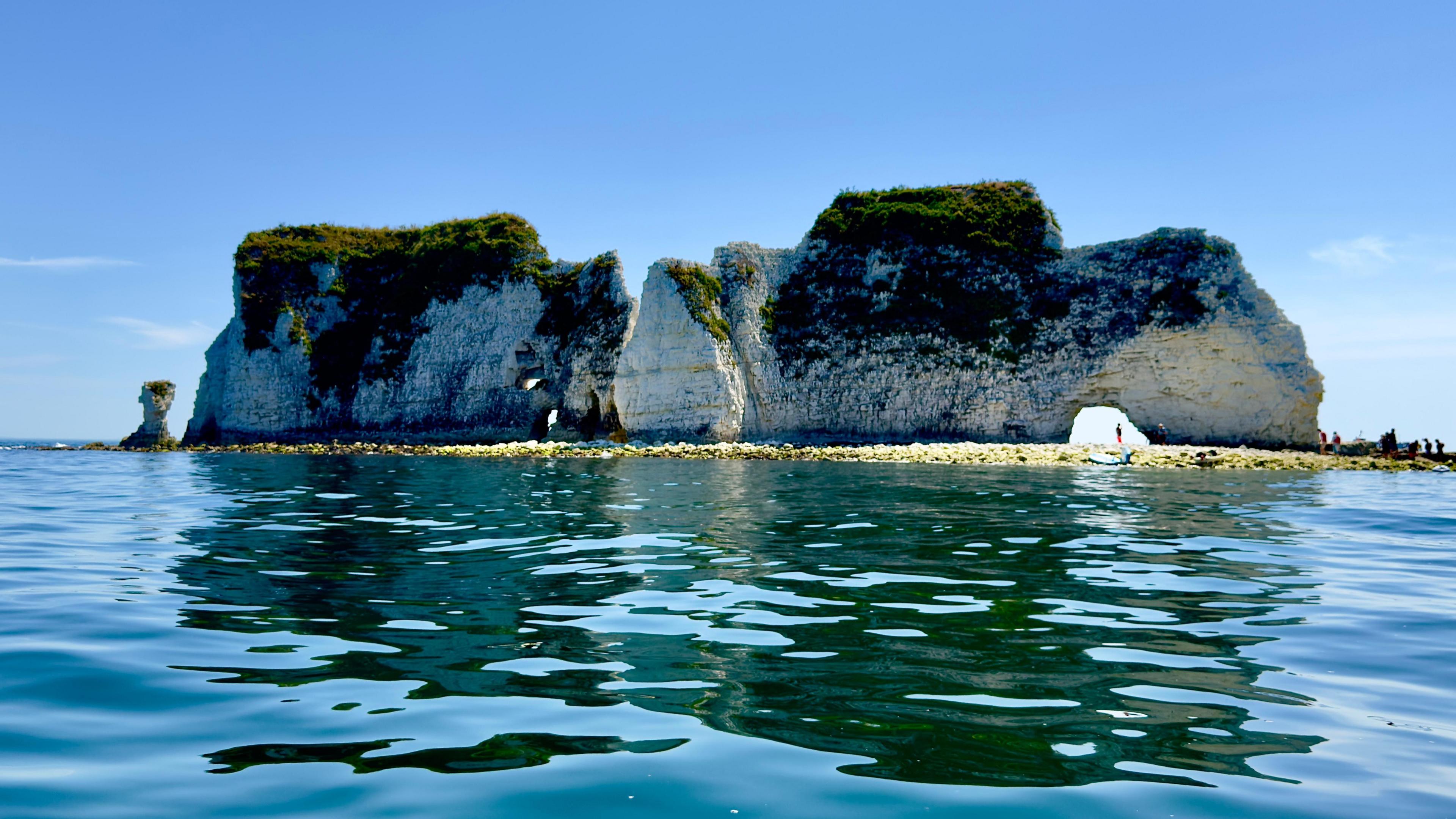 A stoney beach with part of a cliff on it. At the bottom of the cliff there is an arch. A the other end there is a stack. In the foreground is the sea, which looks dark blue.