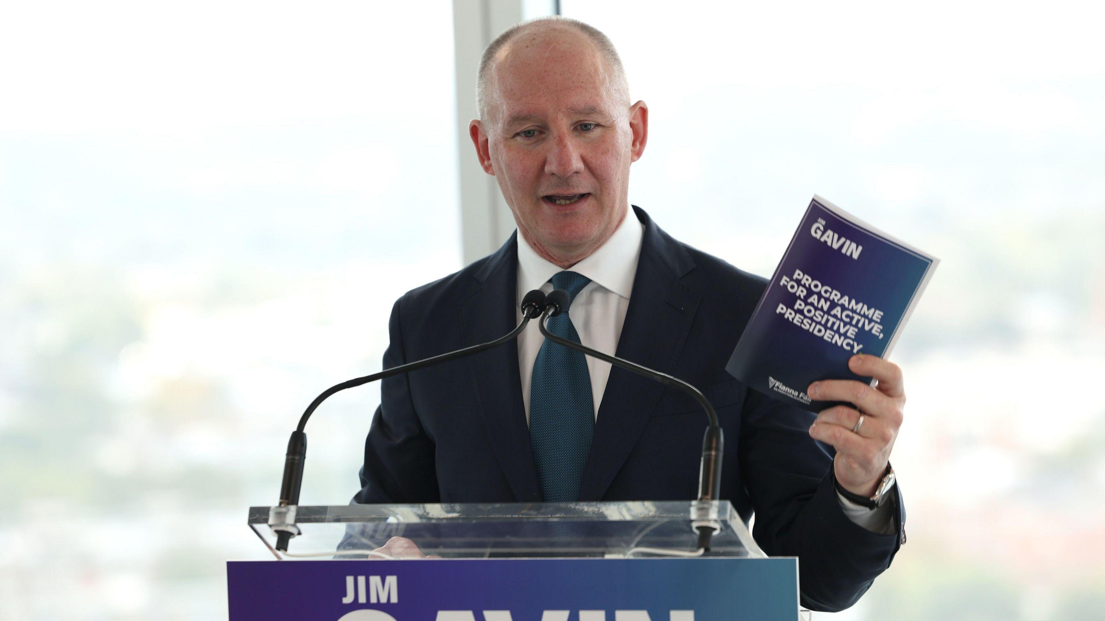 Jim Gavin, standing behind a podium, holding a leaflet in his hand. He is wearing a navy suit, dark green tie and white shirt. There is a large window in the background. 