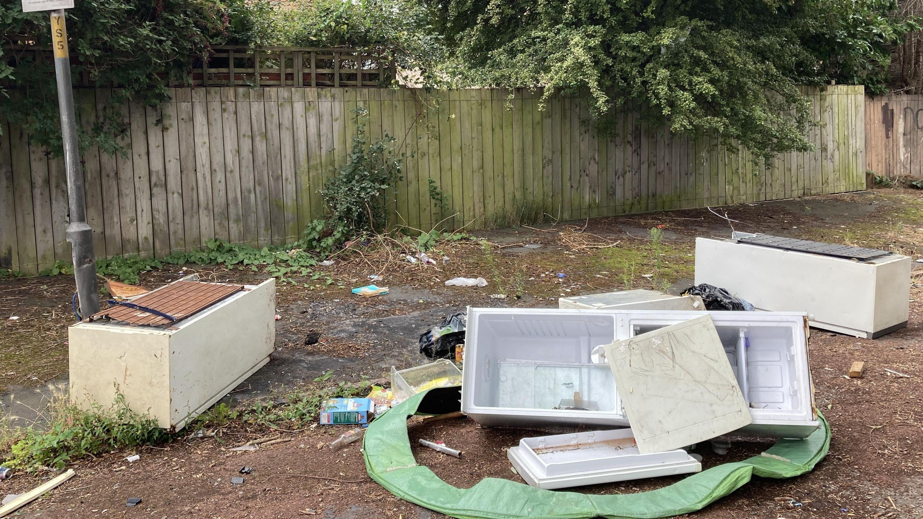 View of the road and pavement with three abandoned fridge freezers - one with the door torn off and its parts and other pieces of rubbish surrounding it. They are in front of a wooden fence, and overhanging shrubbery.