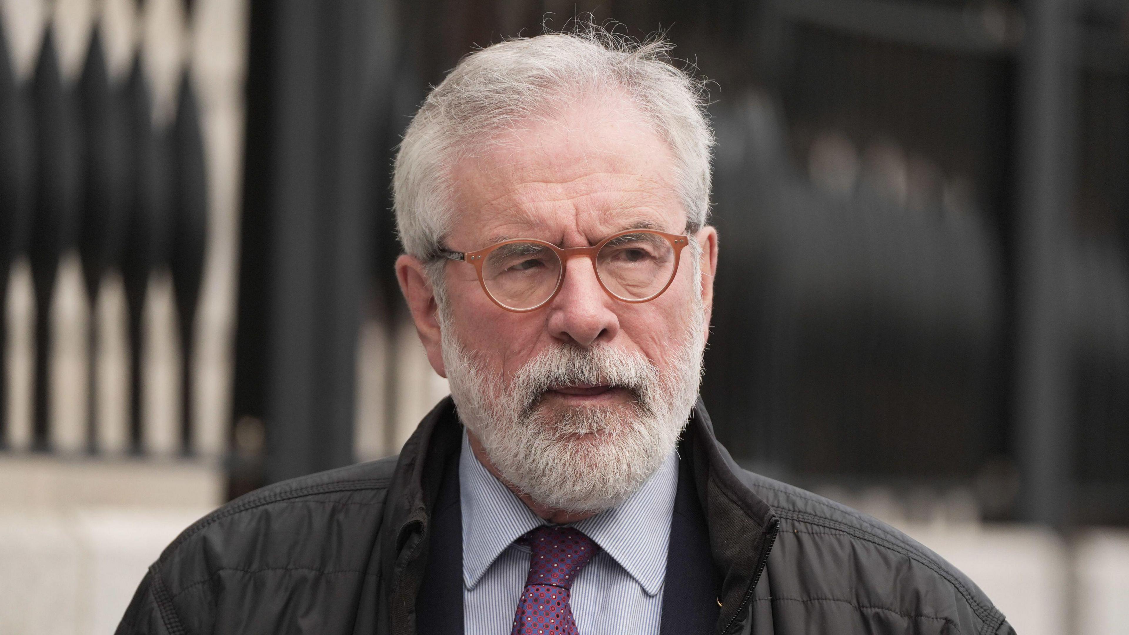 Gerry Adams, with short grey hair and a grey beard. He is wearing a blue shirt, purple tie, dark grey jacket and round orange glasses. The background of the image is blurred.