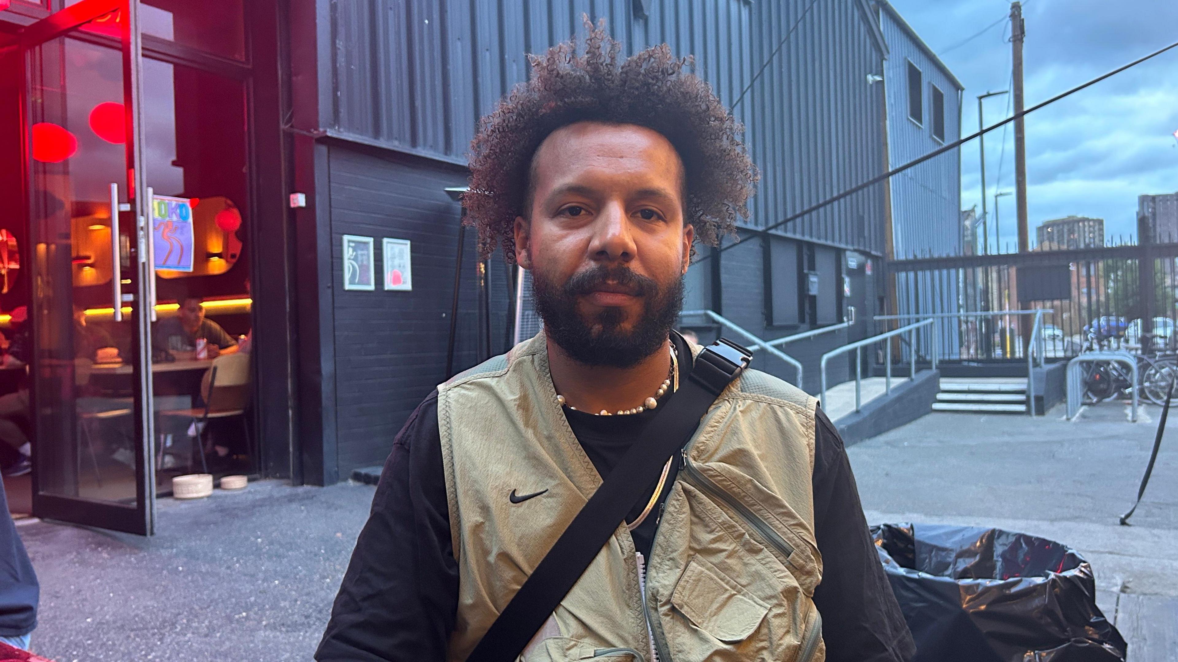 A man sits in the courtyard outside a music venue. He's got afro hair, a beard, and wears a light brown vest/gilet over a long-sleeved black top.