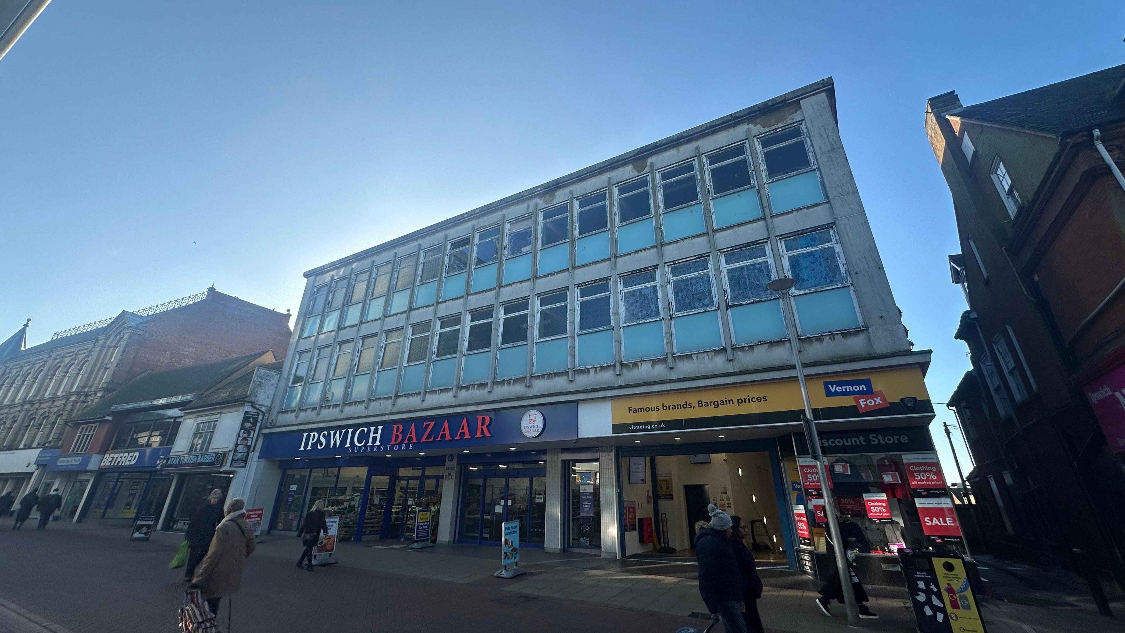 A general view of the building where the flats will be built. People can be seen walking past the building. Shops can be seen on the bottom floor of the building. 