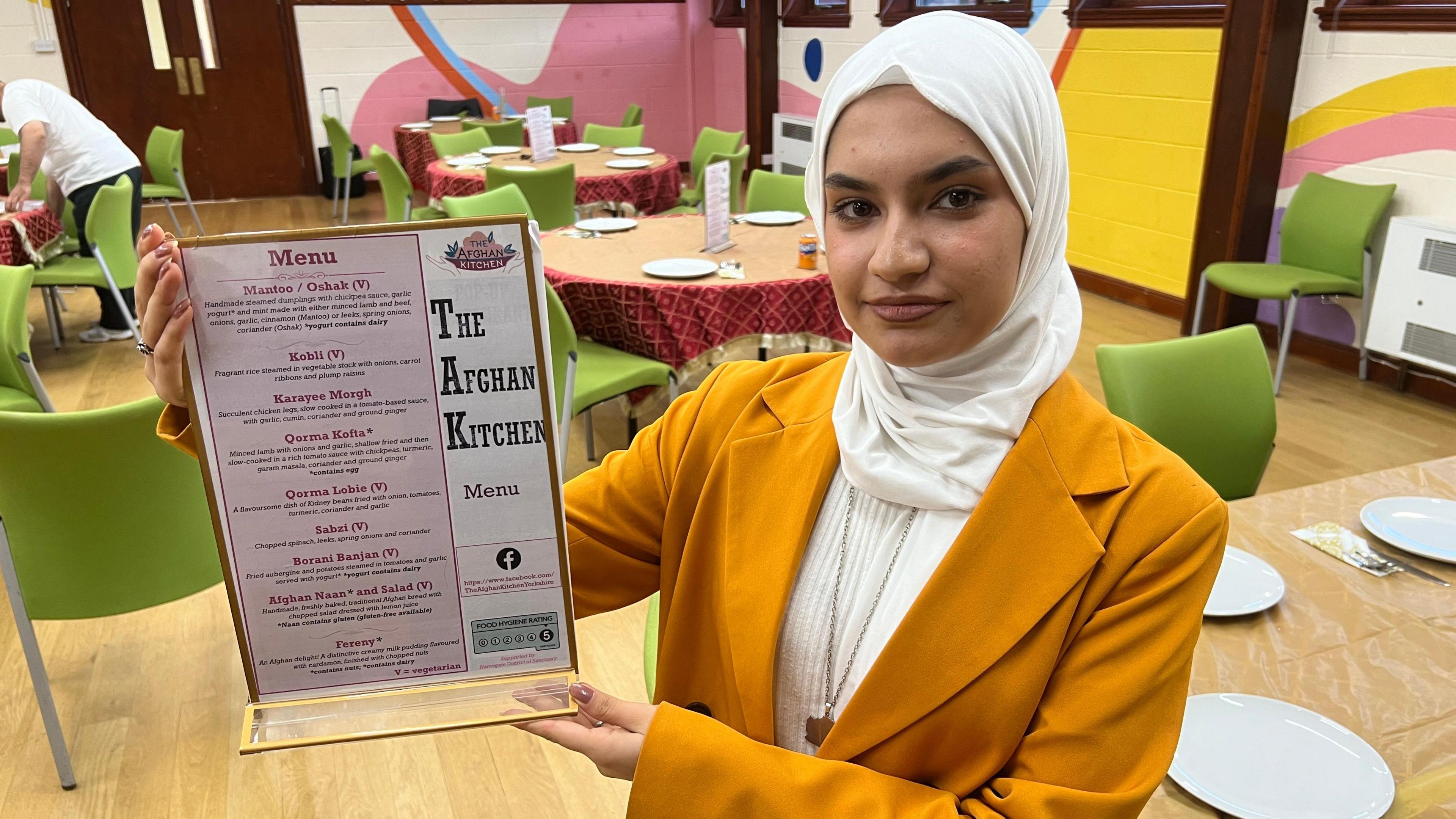A woman dressed in a mustard coloured jacket with a white blouse and head covering. She is holding a clear acrylic stand displaying a printed menu titled 'The Afghan Kitchen. In the background are tables set with white plates and cutlery and green chairs.