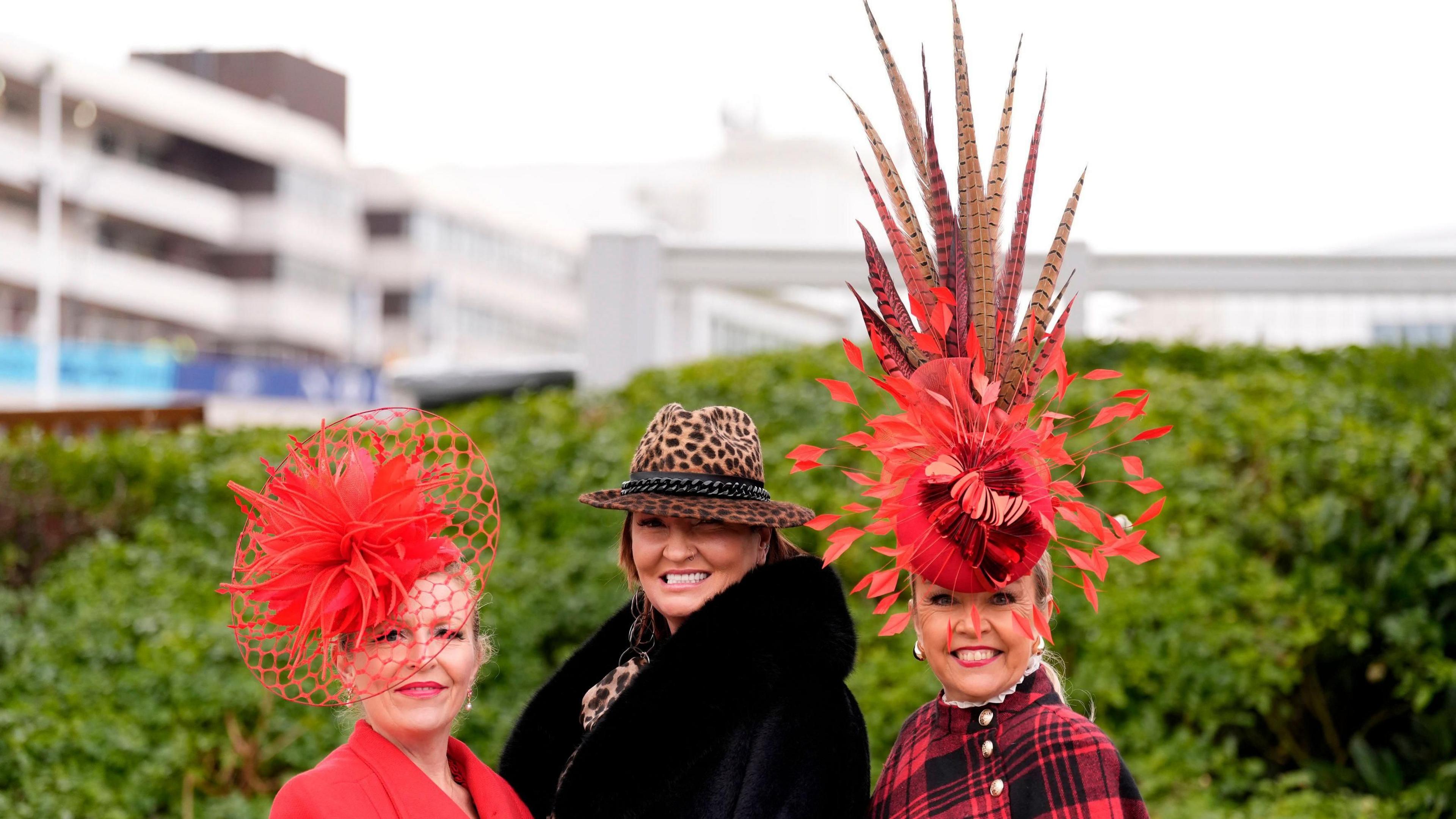 A group of three women dressed in unique hats and bright red headwear at the Cheltenham Festival in front of greenery in the background.