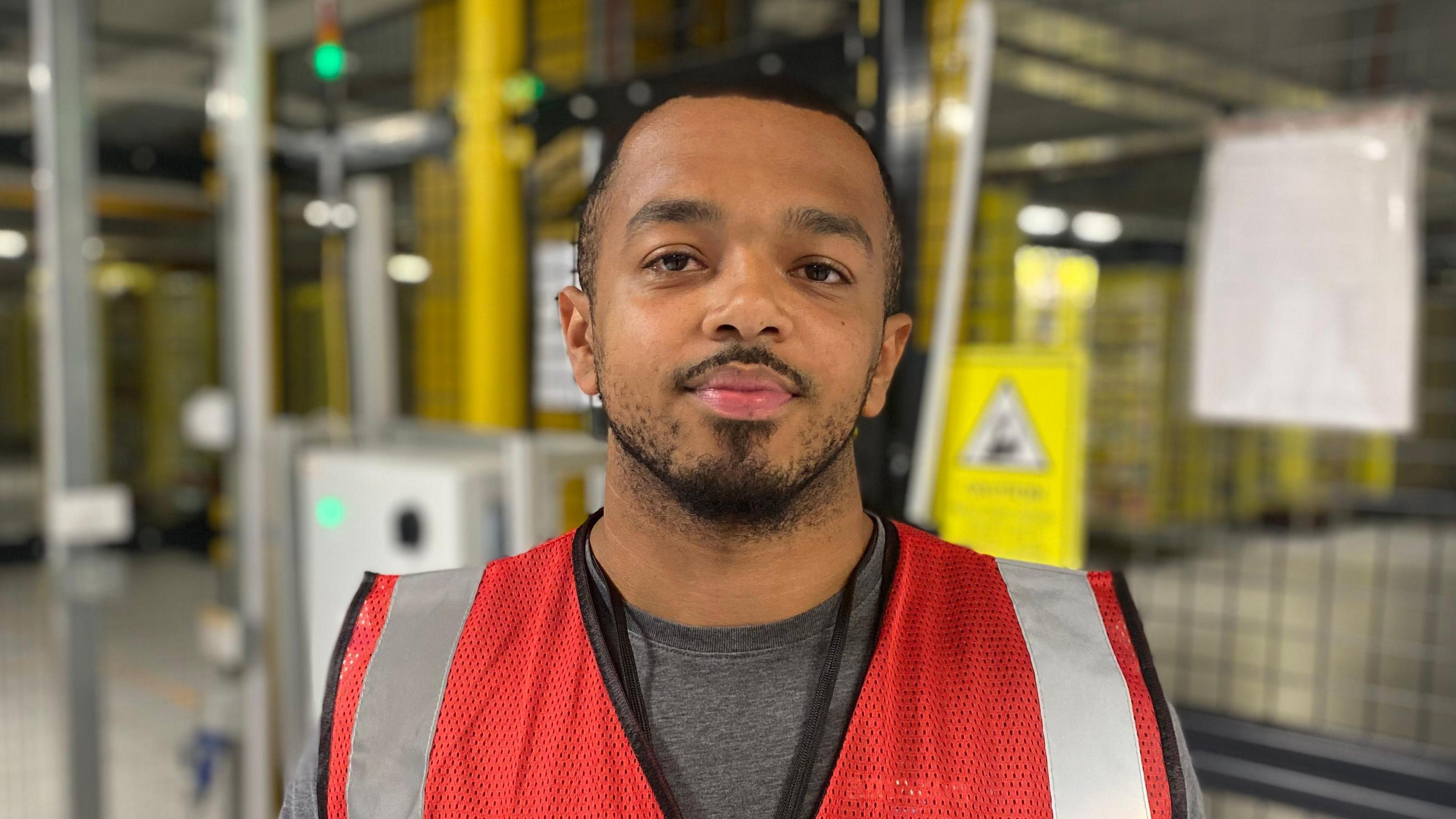 Cole McGrath wearing a red high-vis vest stood in front of a mesh barrier bearing yellow warning signs inside a warehouse.