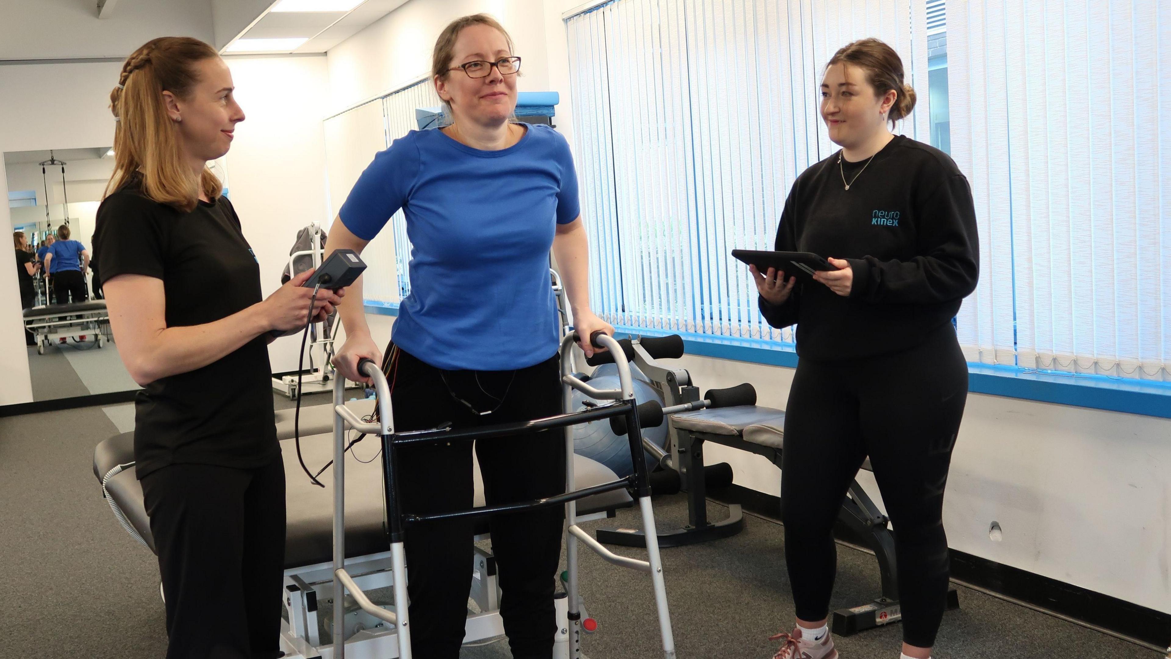 Claire Trivedi, a woman with long blonde hair and glasses wearing a blue T-shirt and black trousers, stands with the aid of a metal walking frame as two members of staff in black stand on either side of her holding monitoring equipment. 