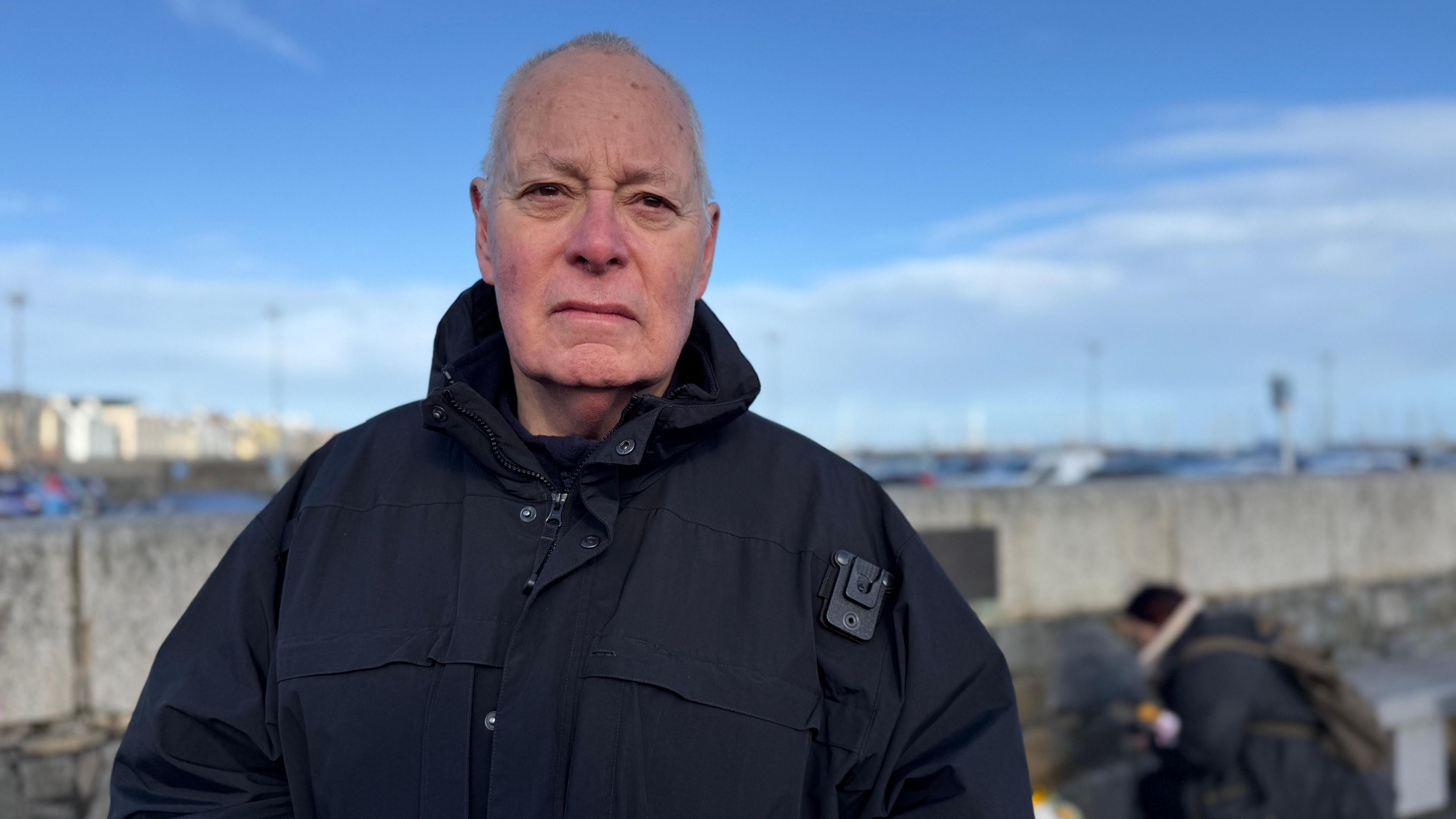 Phil Martin is wearing a black jacket. He is stood is looking into the camera. A blue sky is above him. 