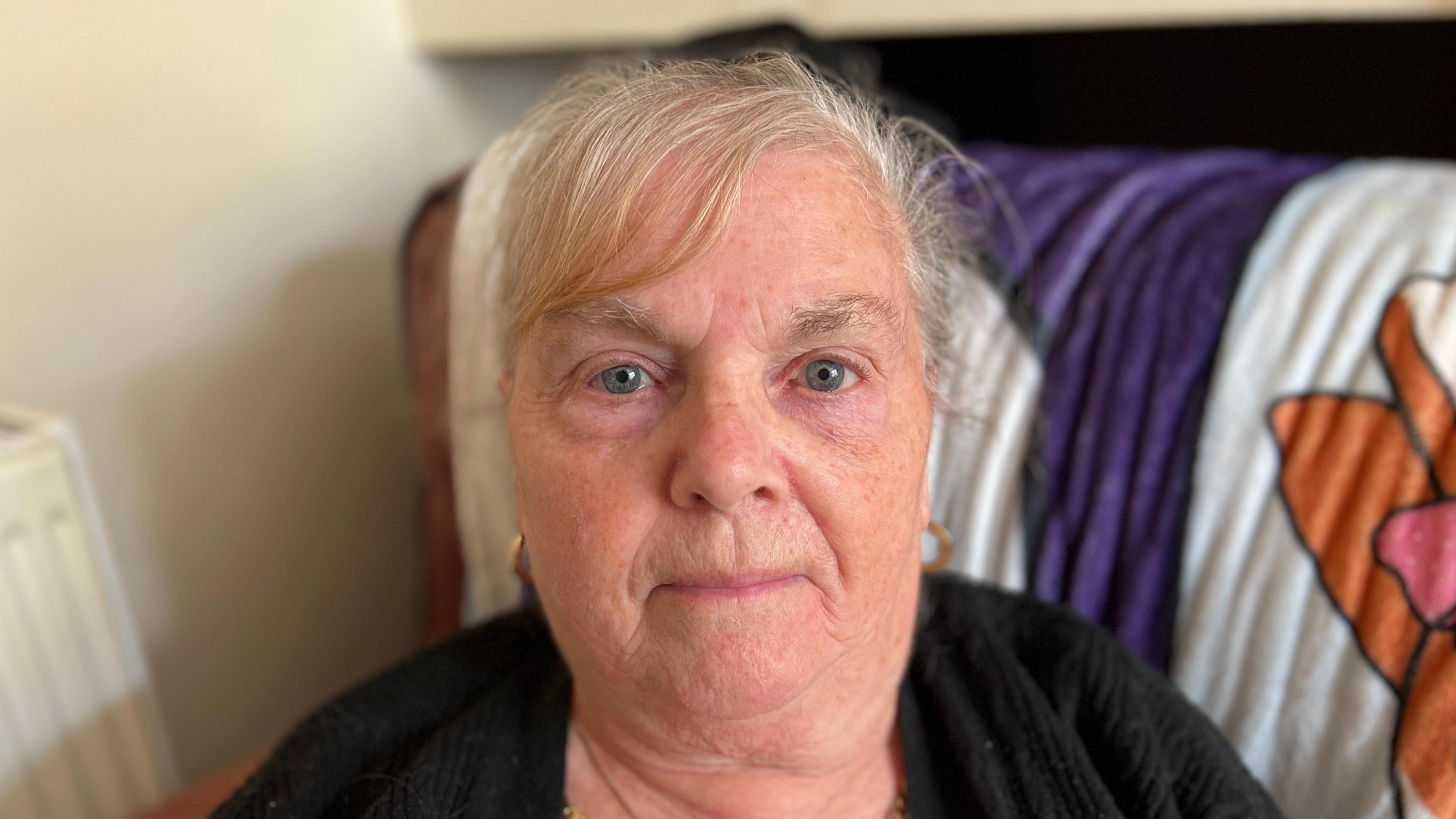 Janet is sitting down, she has blue eyes and blonde and grey hair. She has gold hooped earrings. Behind Janet is a purple, orange and white blanket.