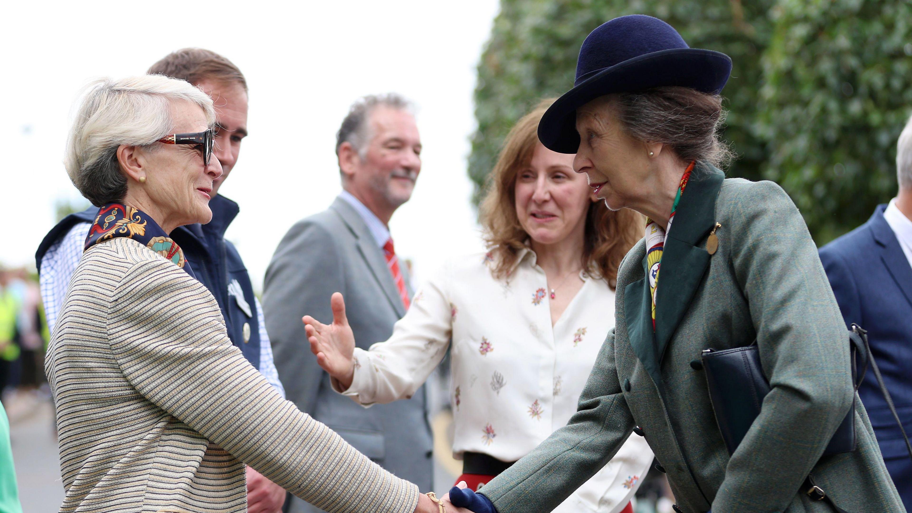 Princess Anne shaking hands with a woman. Princess Anne is wearing a navy hat, with a green coat. 