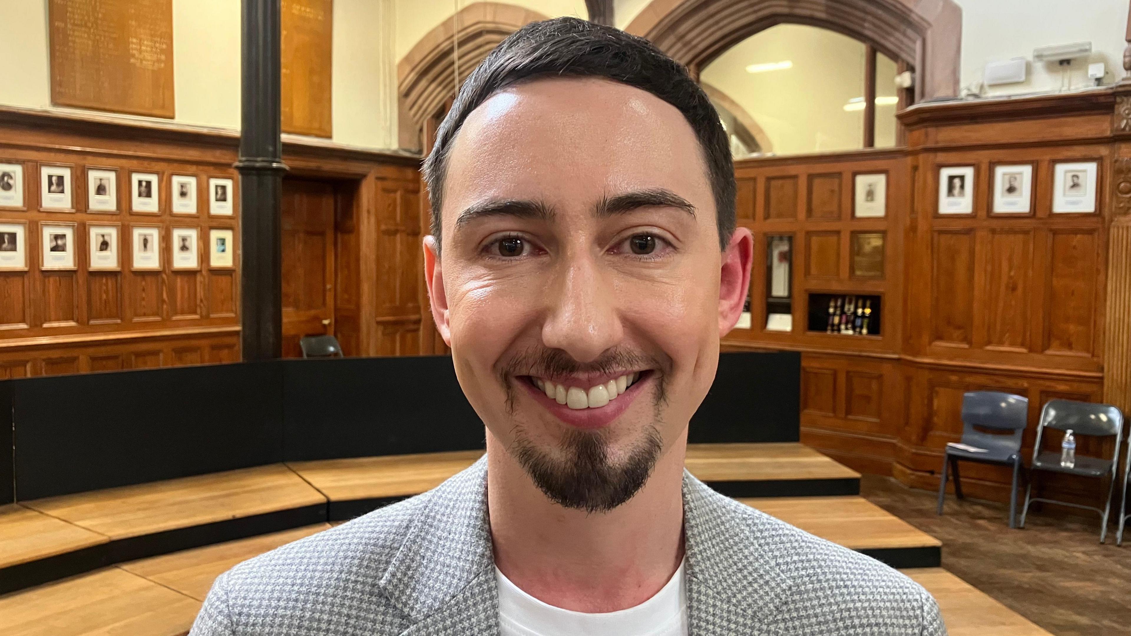 Andrew is standing in a grand looking wooden panelled room with small white portraits in the background. He is smiling at the camera in a grey and white blazer over a plain white t-shirt. He has brown short hair, a goatie beard and brown eyes.