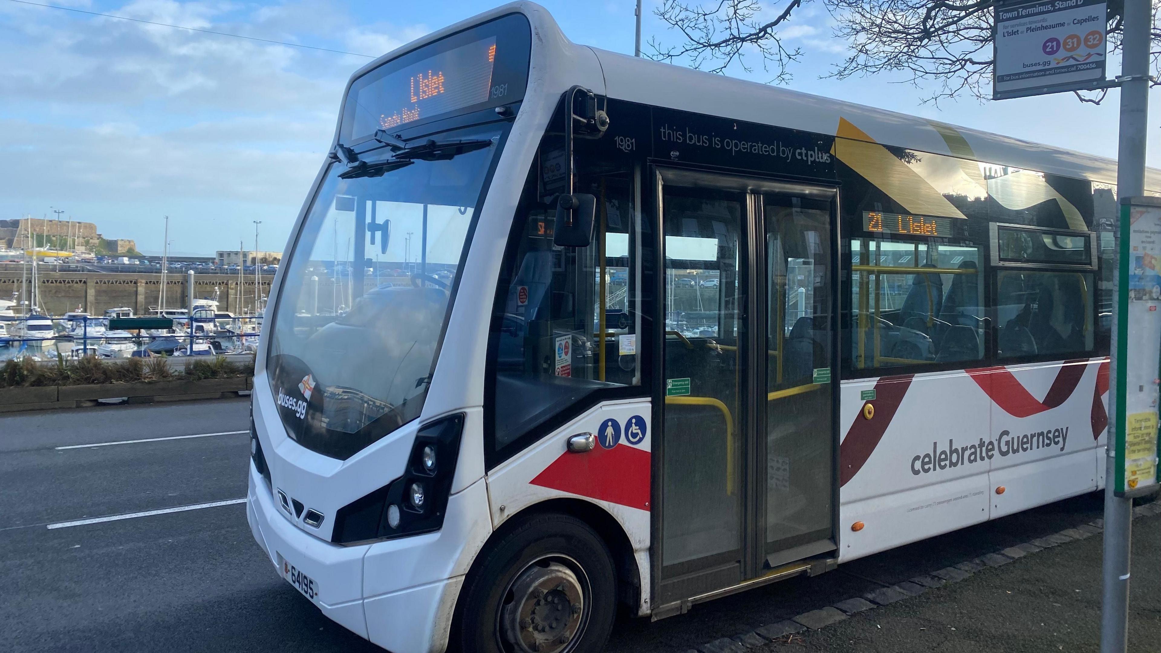 A guernsey bus at the town terminus. The bus is parked at a bus stop. The bus is white with red ribbon decals. The marina is visible behind the bus. 