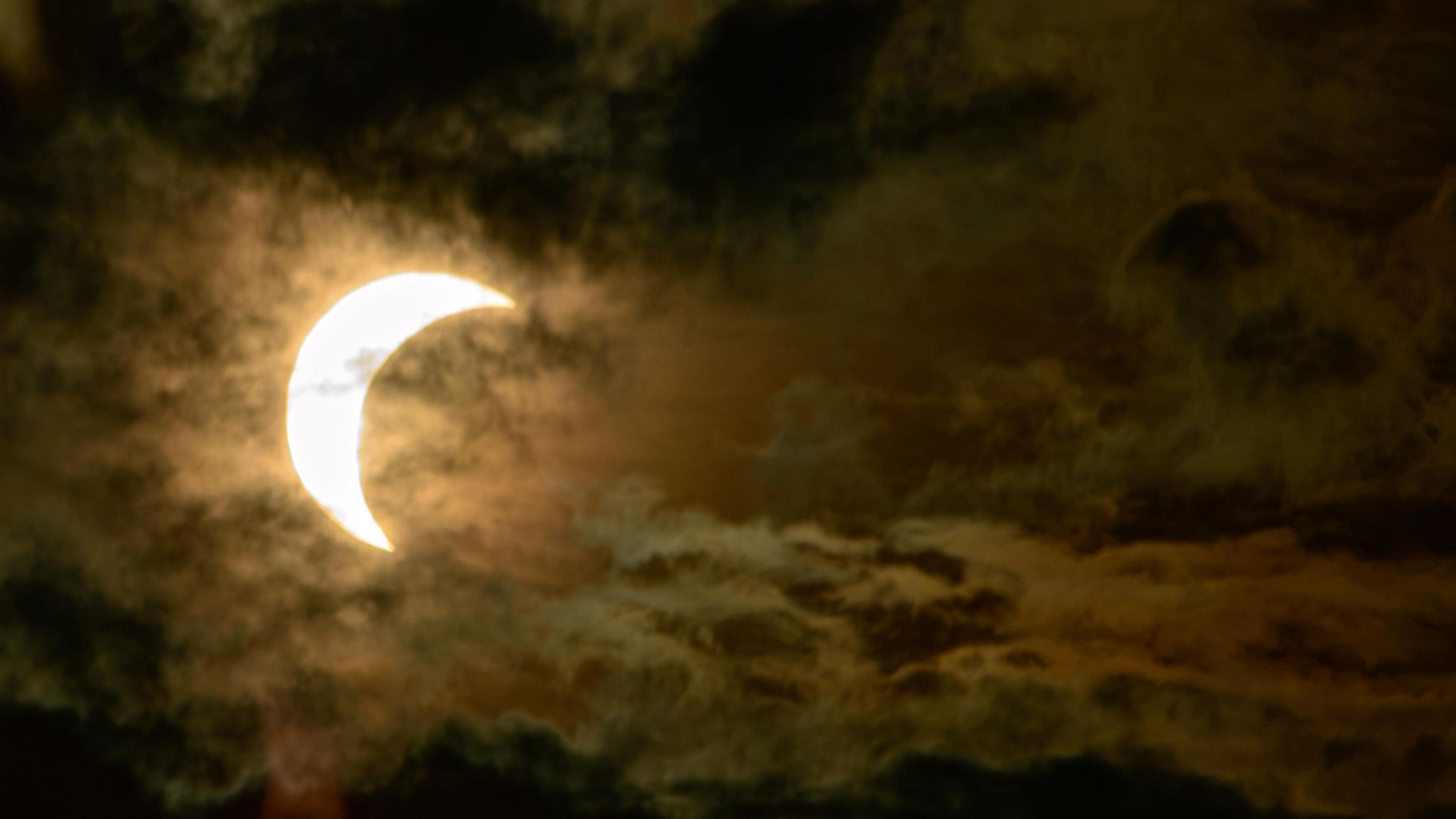 A bright solar crescent is partially obscured by swirling orange-brown clouds. The eclipse is less advanced than in the first image, and the cloud textures are especially prominent across the scene.