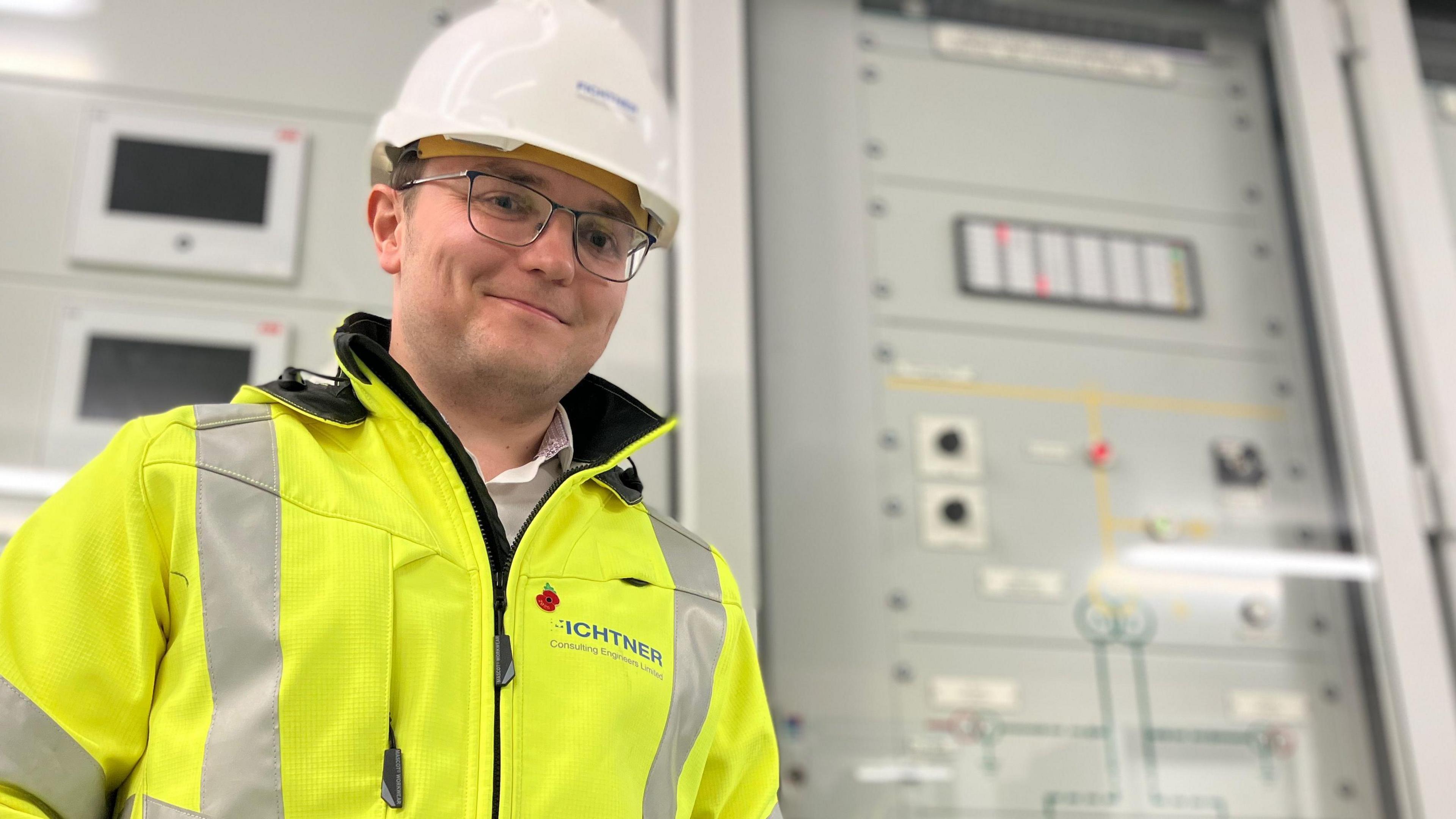 A man in glasses, wearing a 'high-vis' yellow jacket, white hard-hat and gloves smiles at the camera. Behind him in cabinets is grey electrical equipment.