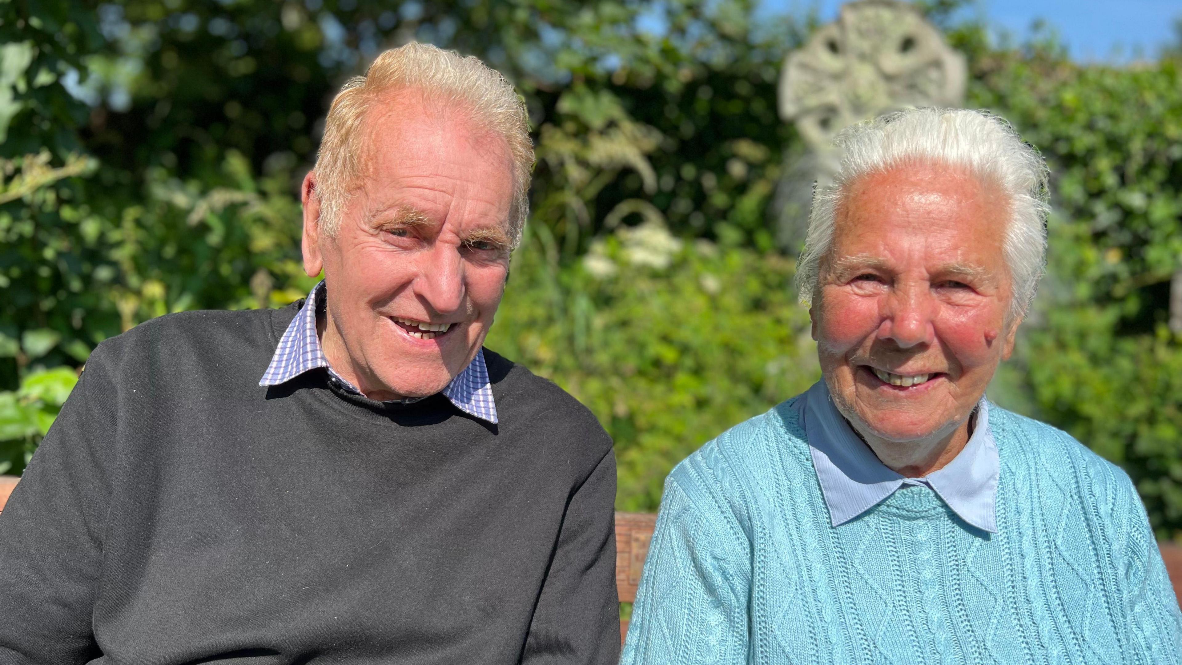 An elderly man in a dark jumper and an elderly woman in a bright blue jumper sit in the sunshine in a churchyard in Illogan