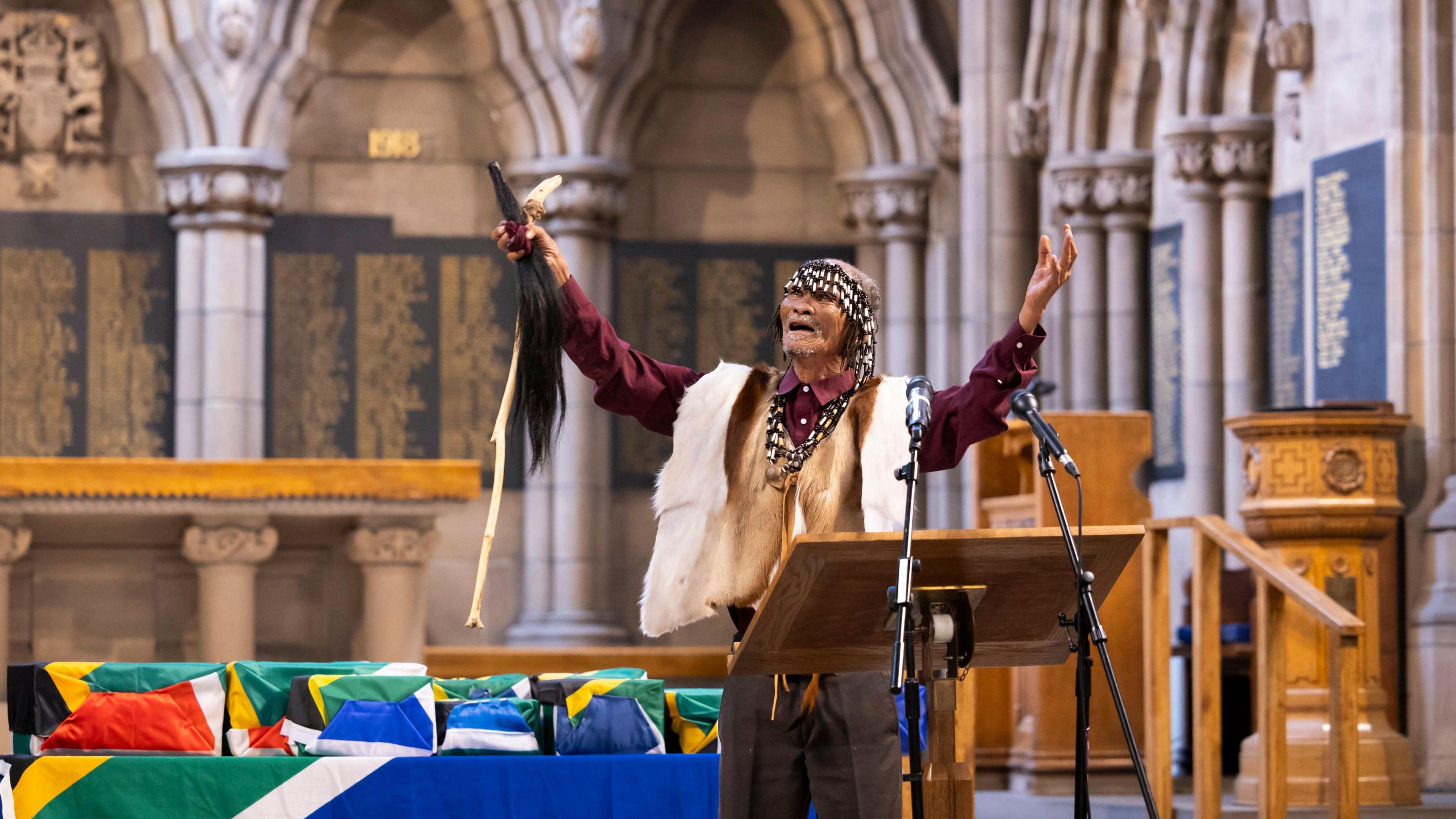 A man standing in a chapel, behind a lectern and with microphones. He has his arms raised, as if appealing to a higher power. Behind are several small coffins or boxes, containing human remains of his ancestors.