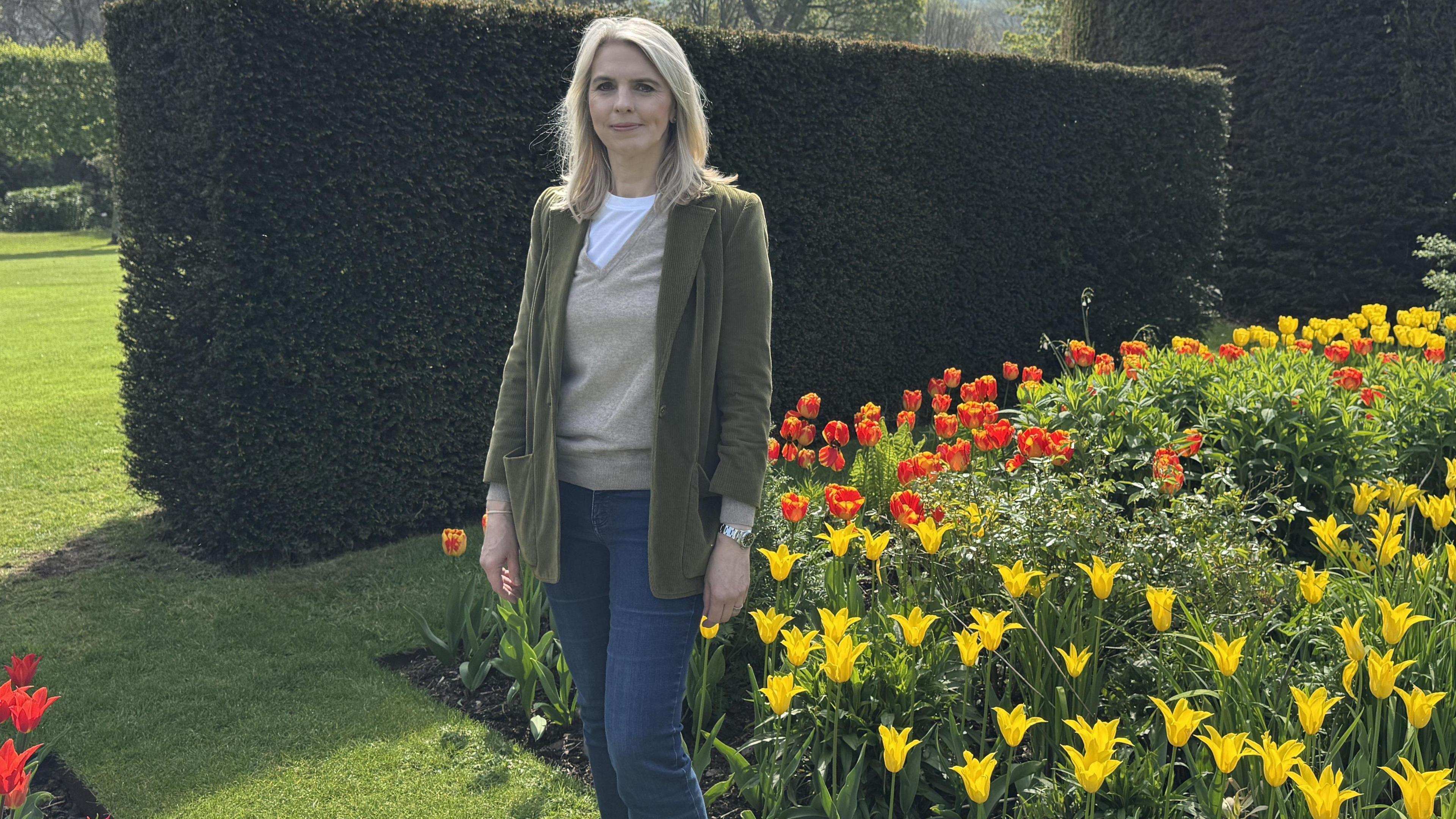 Lady Antrim wearing a green jacket, cream jumper and white top. She has blue jeans and has blonde hair. She is surrounded by tulips that are yellow and orange and shrubbery