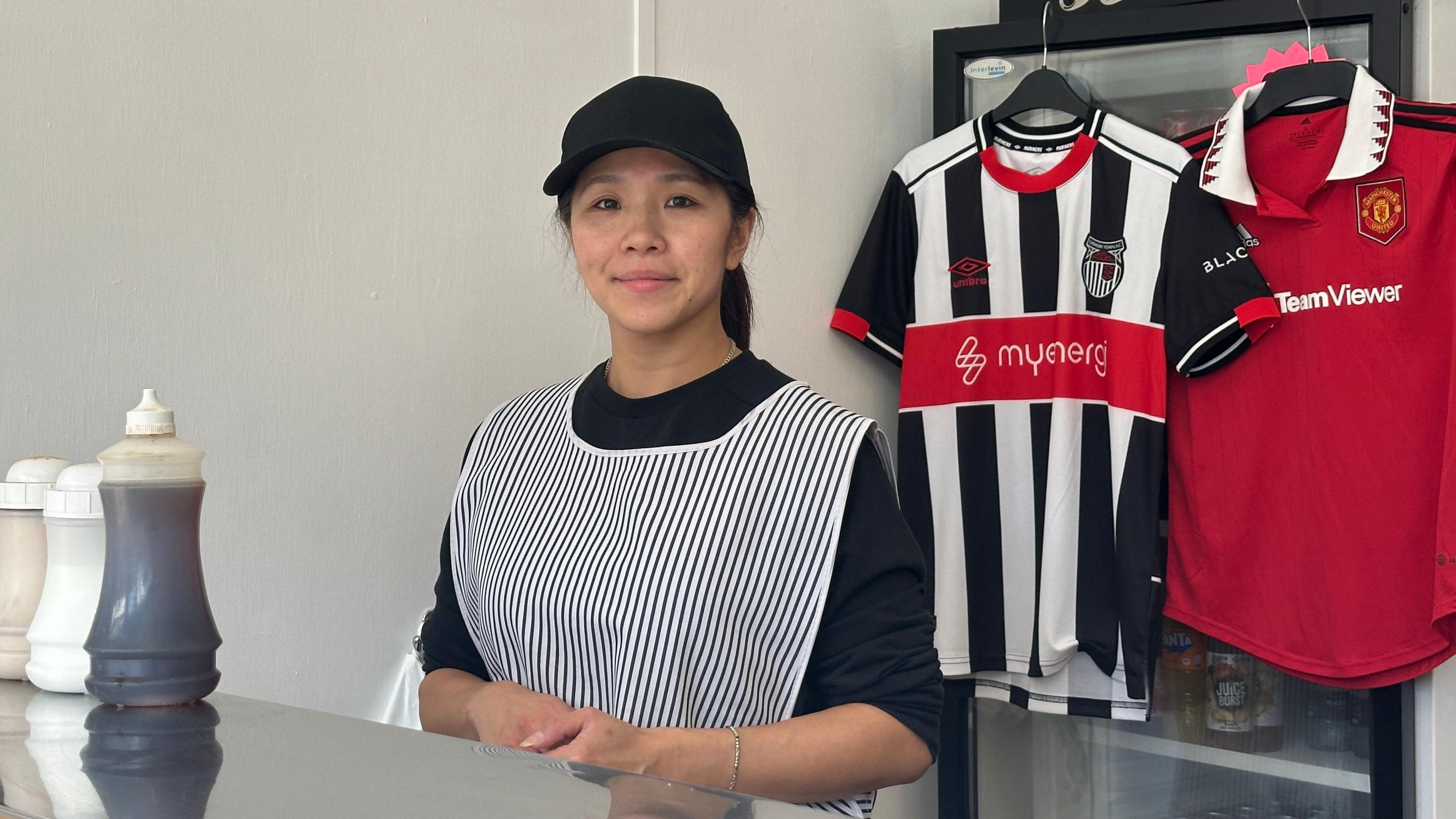 A woman wearing a black cap and black pinstriped serving pinafore stands behind a metallic, reflective serving shelf. A bottle of vinegar and salt and pepper pots stand on the shelf. Grimsby Town and Manchester United shirts are hung up in the background.