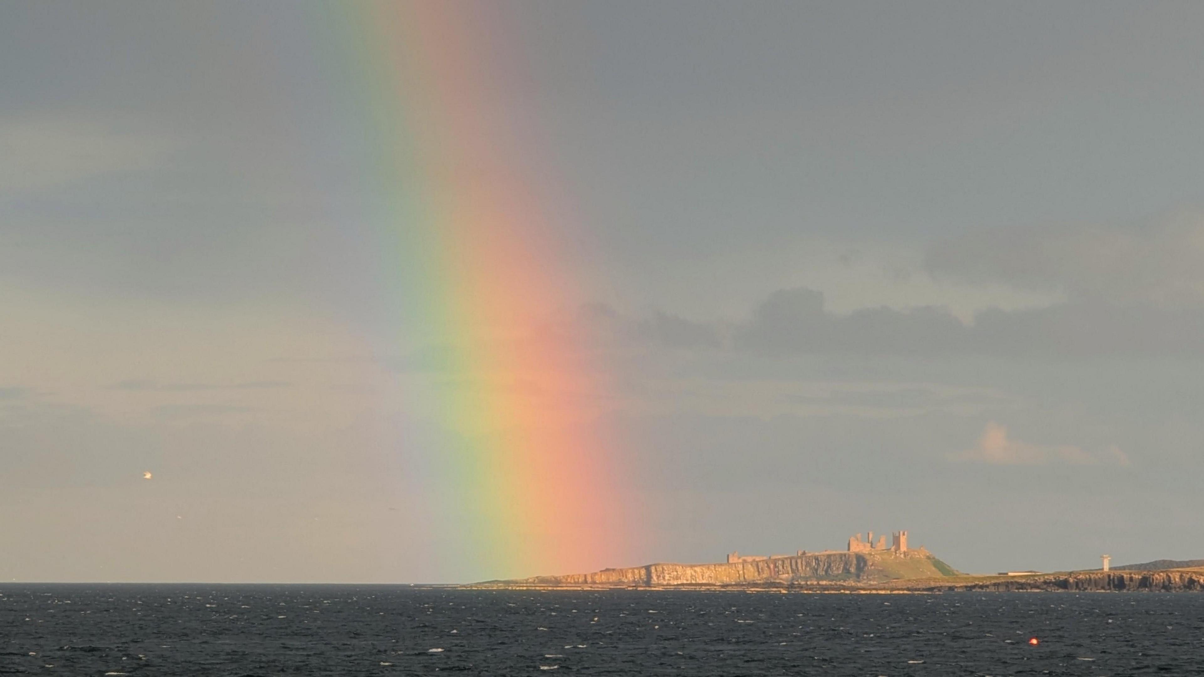 The end of a rainbow comes down over the Northumberland coast. Castle ruins sit on the cliff side.