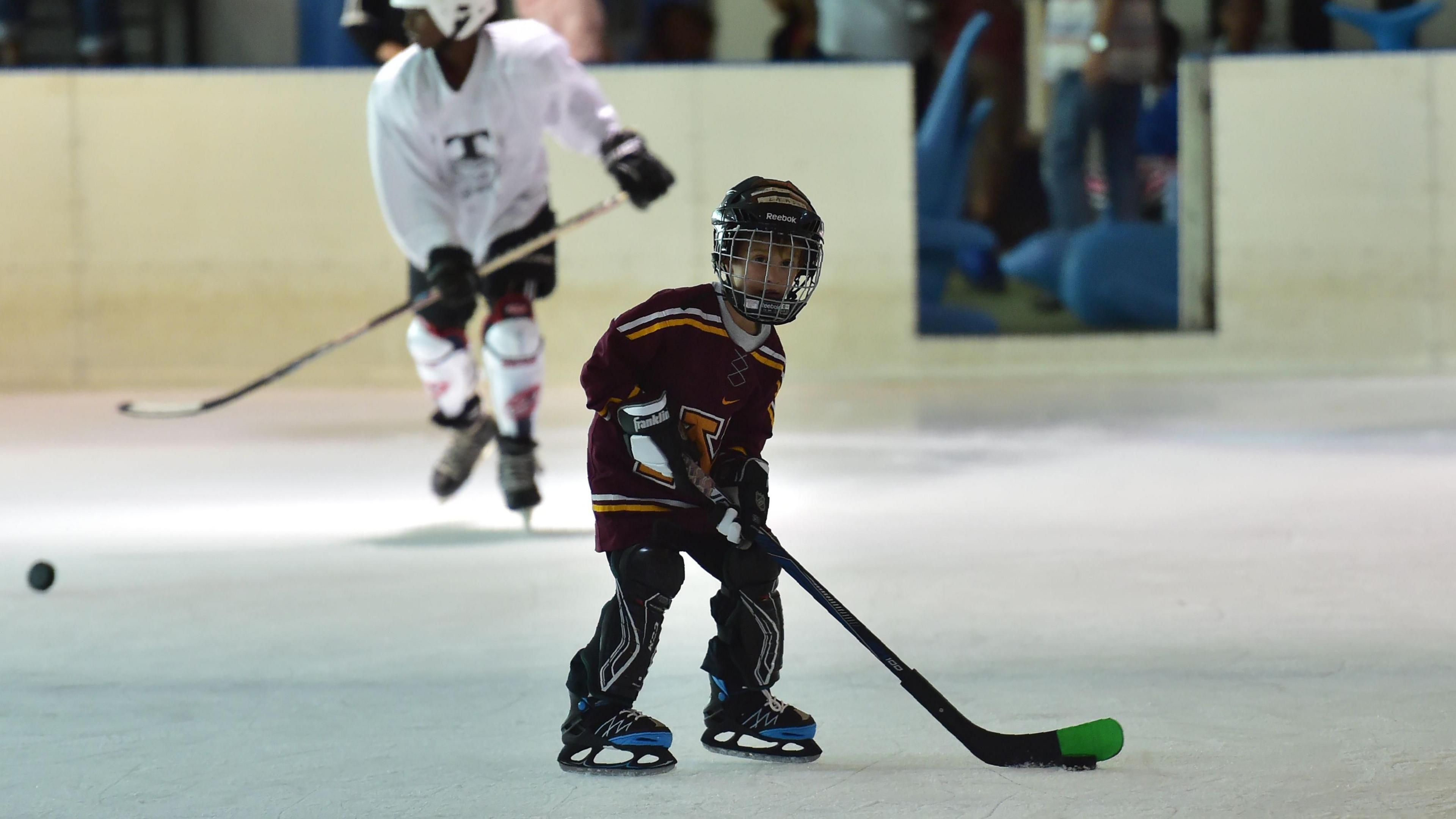 A child, wearing predominantly black ice hockey gear, controls a puck on an ice rink while another player is seen out of focus wearing white in the background