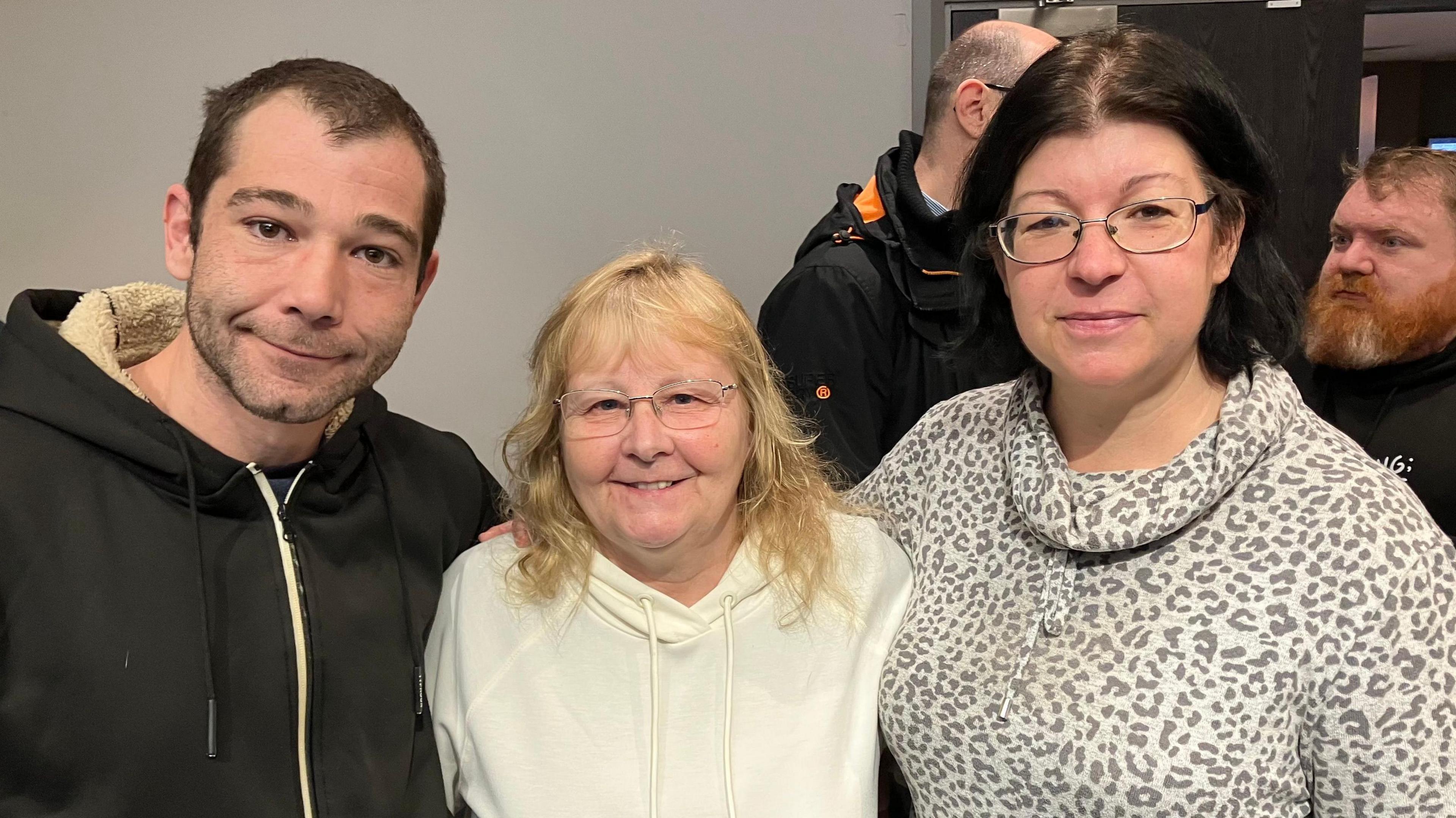 Three local people, two women one man, with arms around each other smiling. Both women are wearing glasses and jumpers. The man is wearing a black hoodie.
