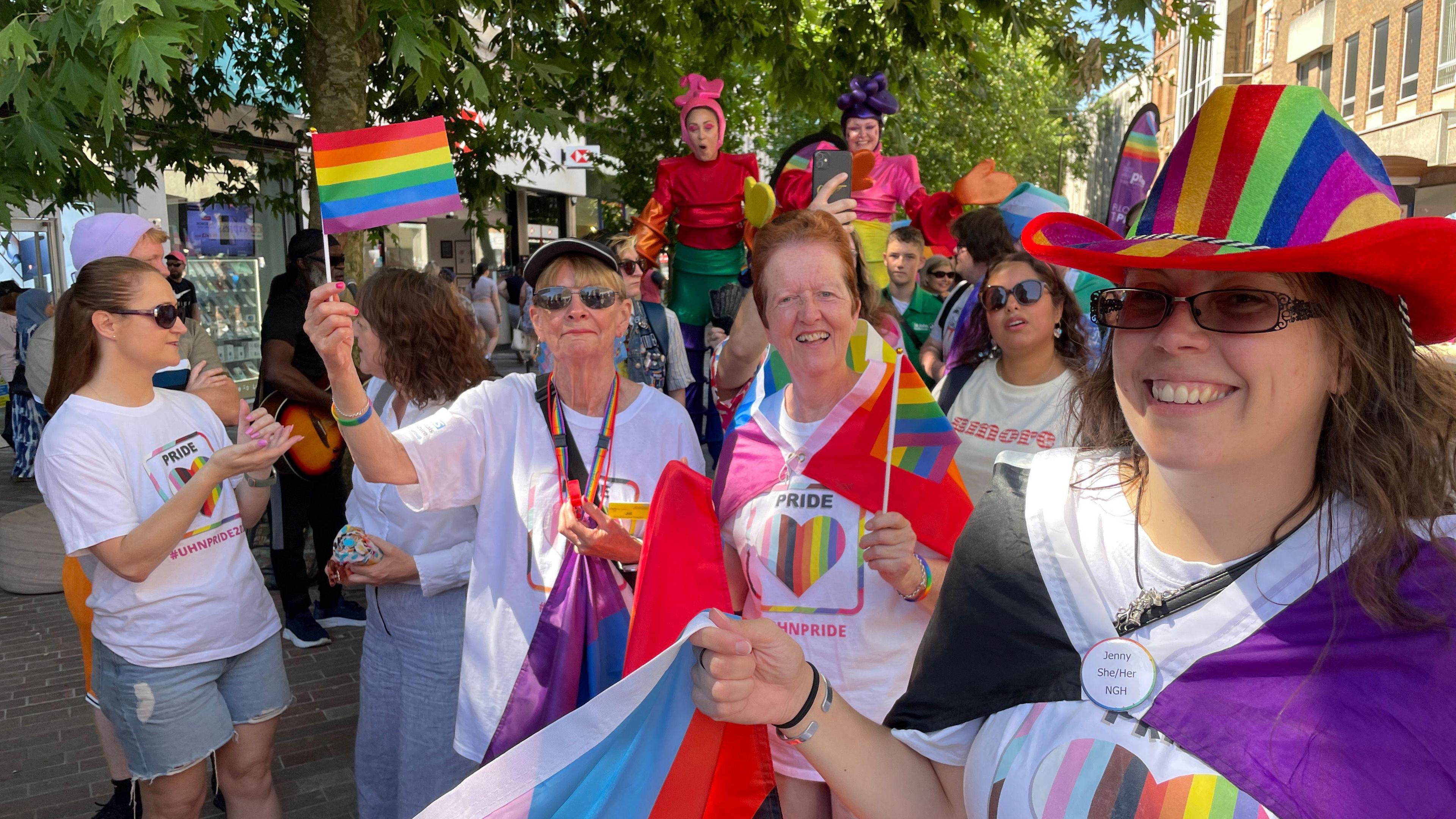 A group of women holding small pride flags on white sticks. They also have flags wrapped around their shoulders. They are walking through the town as part of the parade and crowds of people can also be seen behind them. 