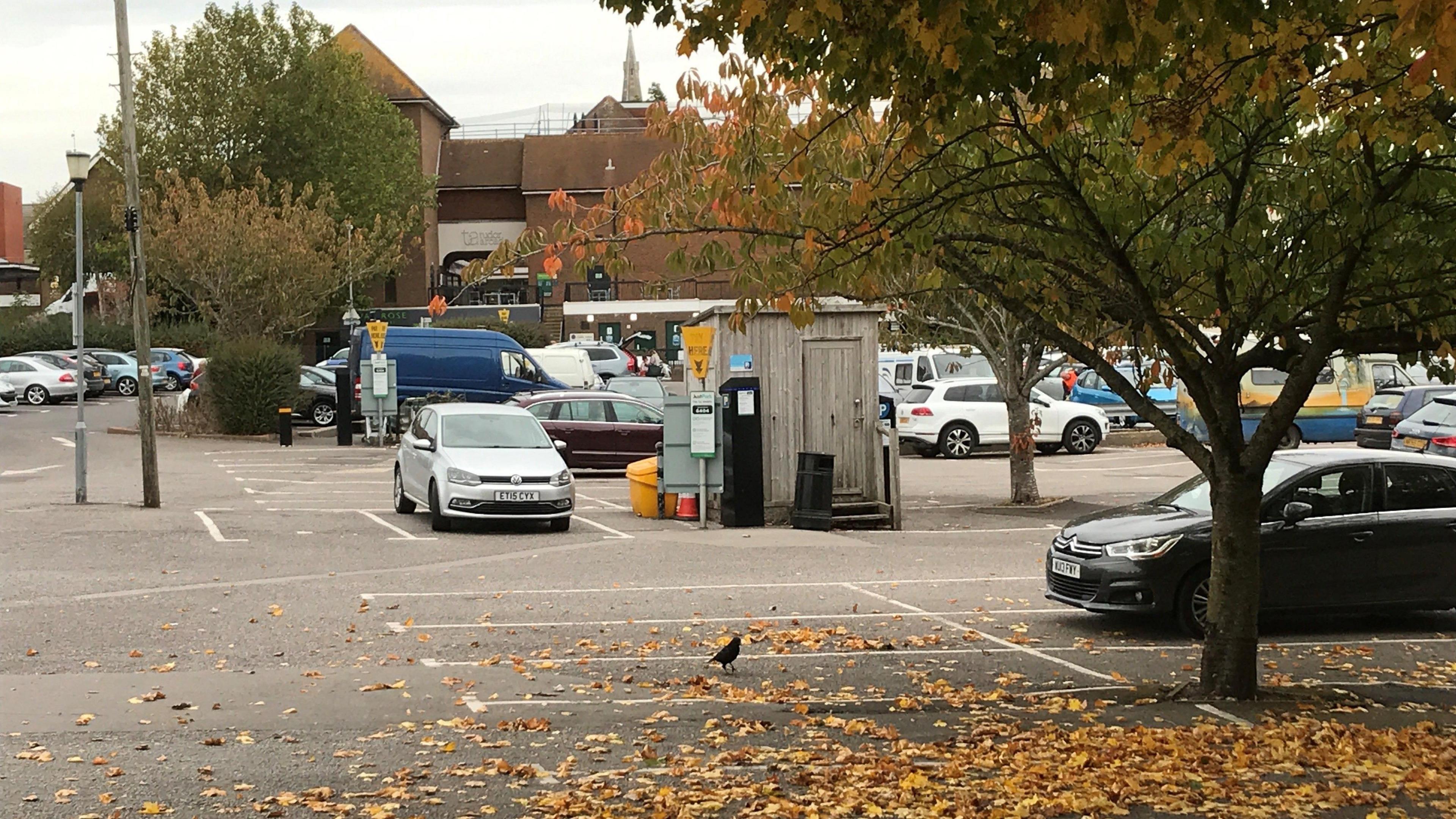 A car park with lots of leaves on the ground and a few cars parked in it. The sky is grey. The car park is in Dorchester.