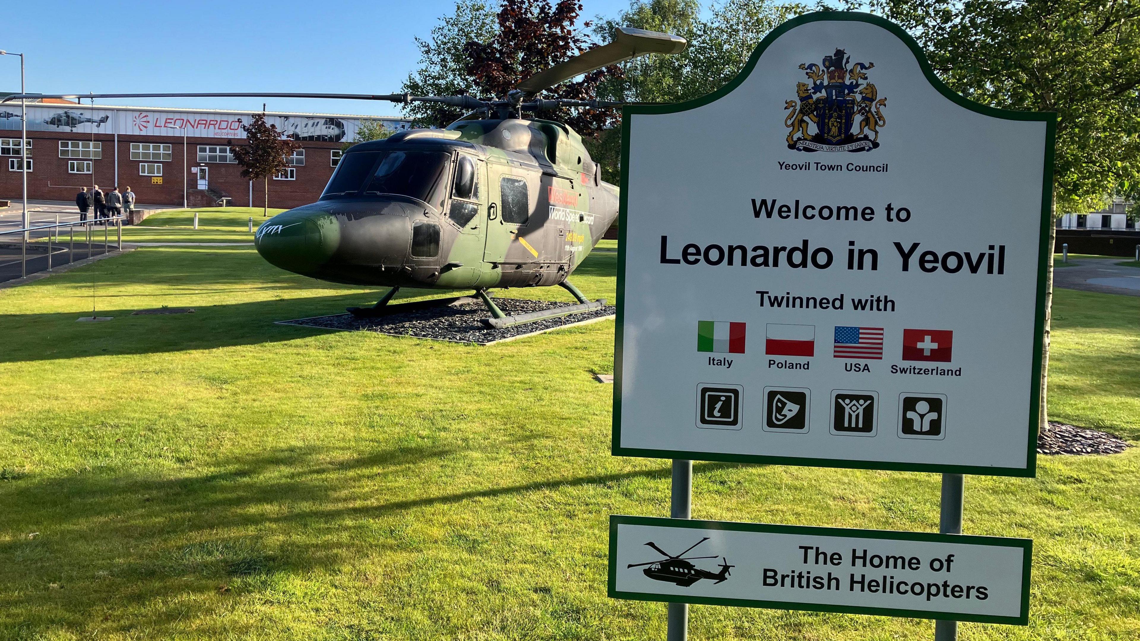 Leonardo Helicopters site in Yeovil. A helicopter can be seen on the grass and a sign reading "welcome to Leonardo in Yeovil" is in the foreground of the picture.