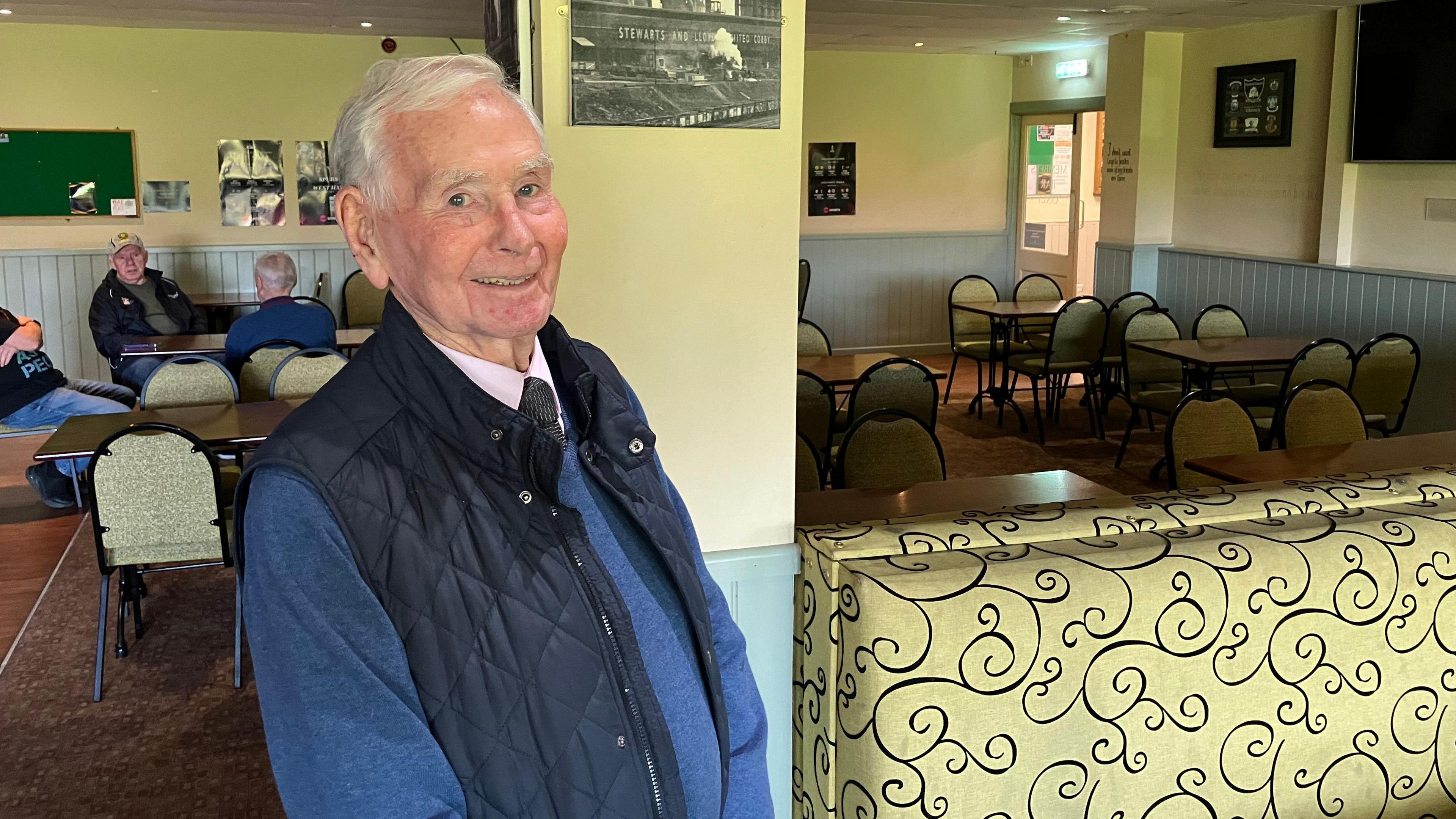 Frank is standing in bar area of the Irish centre and smiling directly at the camera. He has grey hair and wears a blue jumper, a navy gillet and a shirt and tie. He is next to a pillar with several tables and chairs in the background.