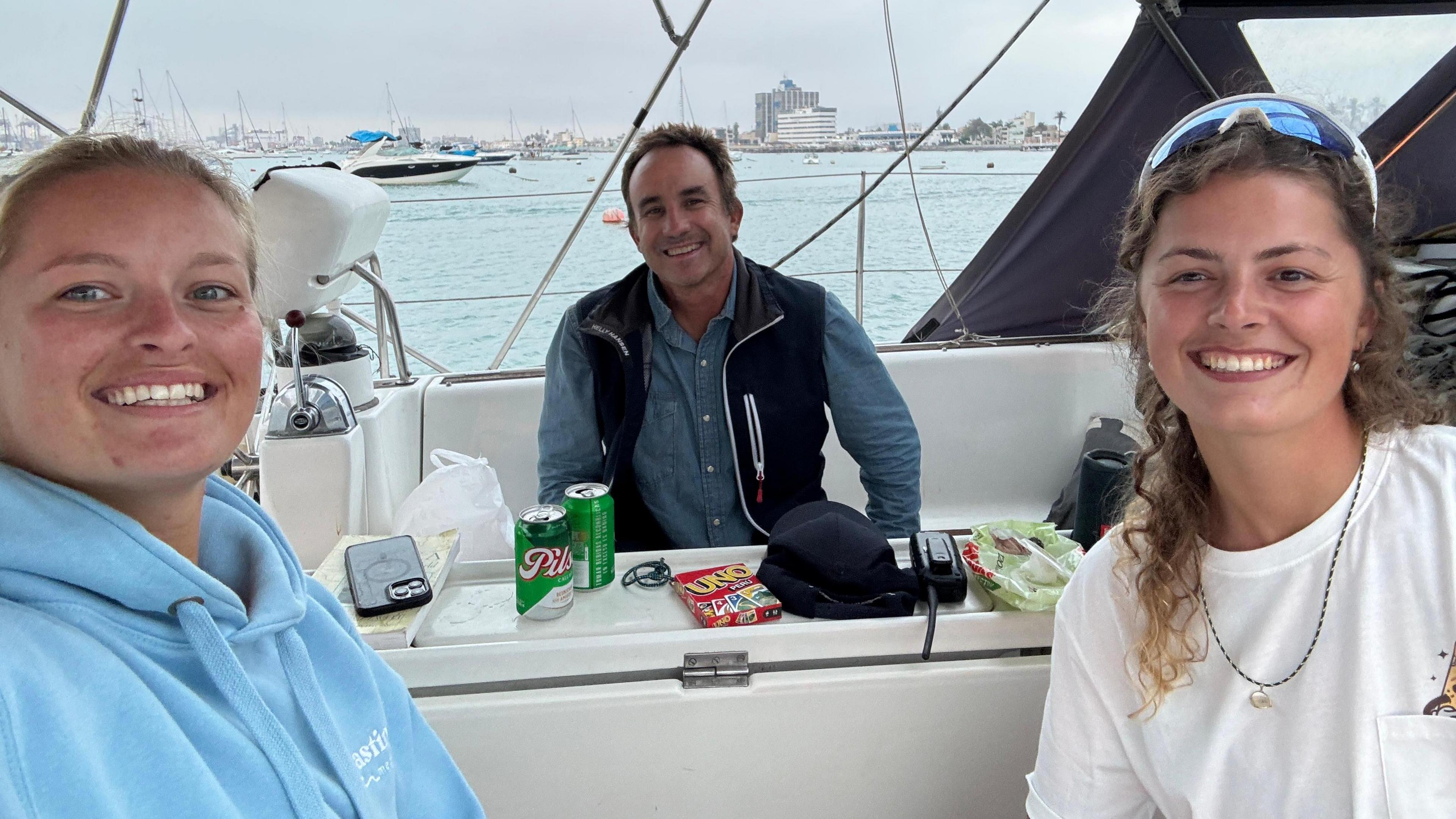 Jess Rowe is on the left of the photo, wearing a blue hoodie with her blonde hair tied back. Miriam Payne is to the right, wearing a white T-shirt and sunglasses on top of her head. She has brown curly hair. They are sitting on a boat, with sailor Alec Hughes sitting on a bench behind them. He has short brown hair and wears a long-sleeved blue shirt and navy gilet. All three are looking at the camera and smiling.