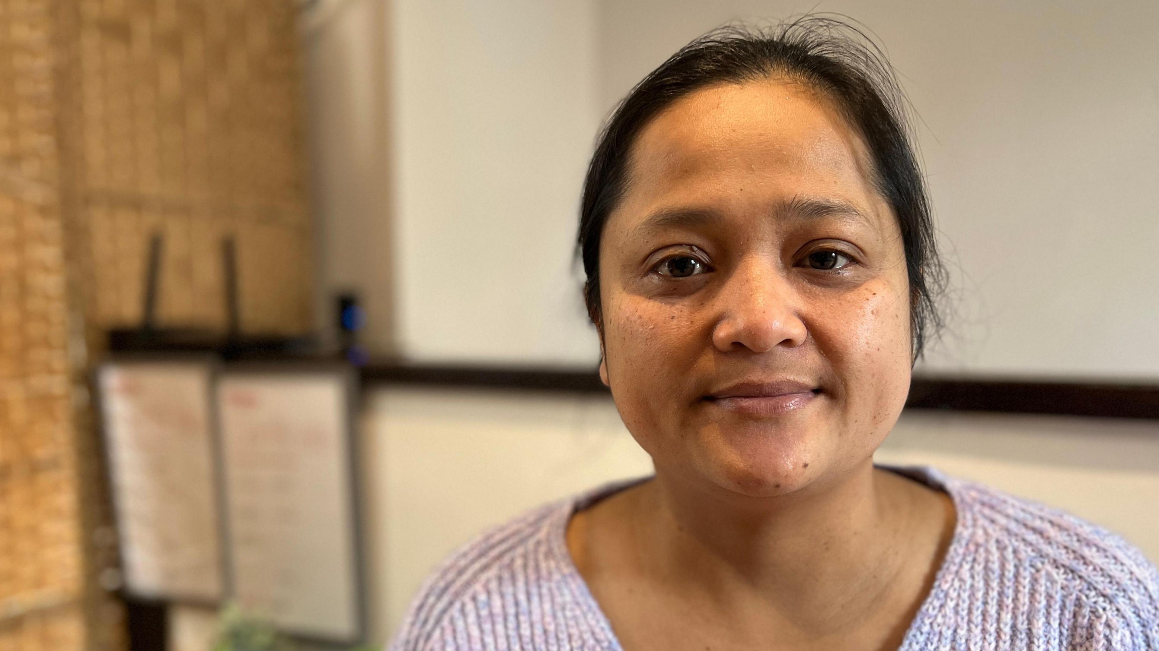 A woman with brown hair, tied back. She is wearing a light-purple cardigan and has brown eyes. The background is out of focus but shows the interior of a shop.