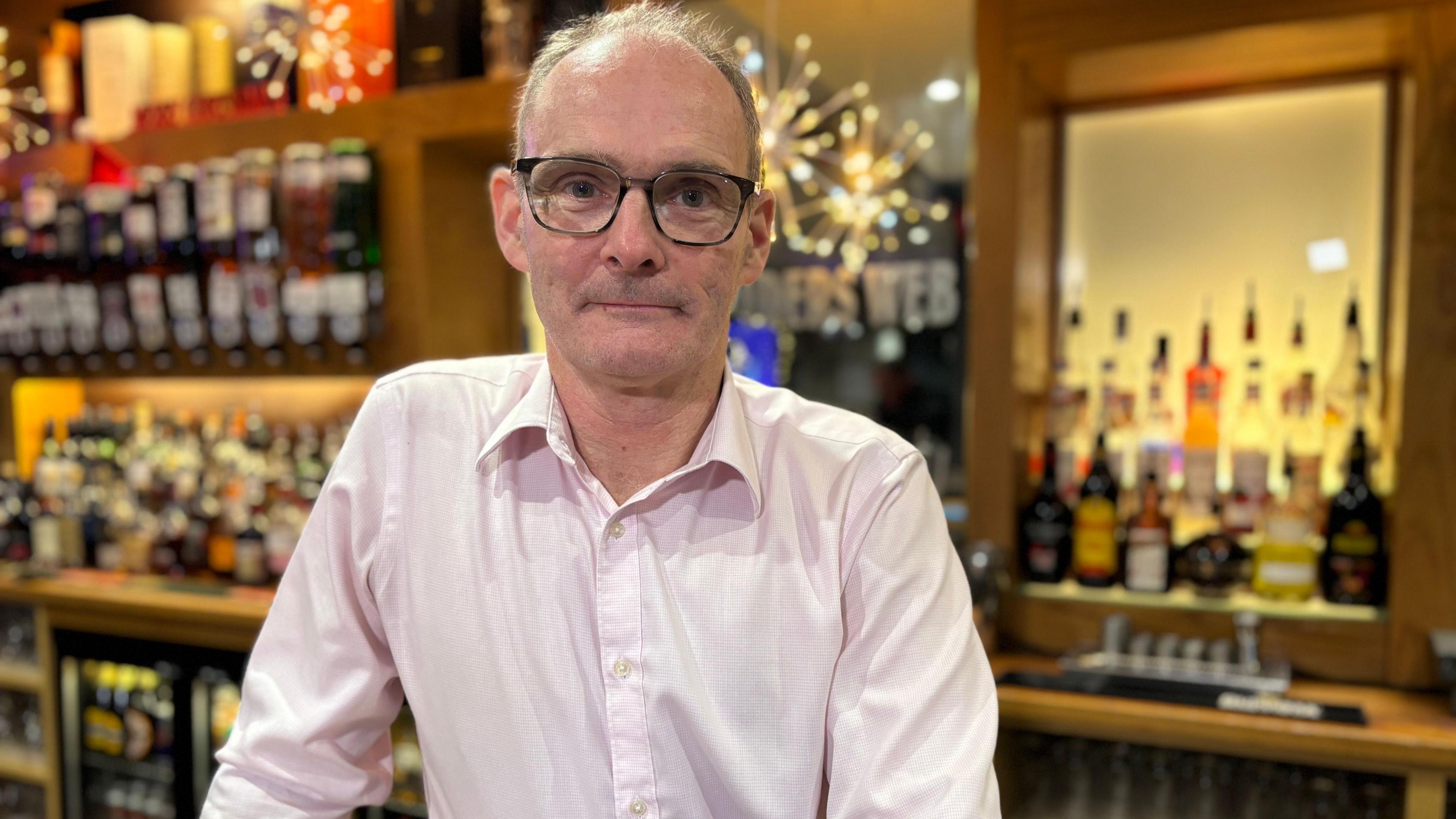 Mark Milne is a balding man with black-rimmed plastic spectacles and a pink open-necked shirt. He is standing in front of a lit bar gantry full of bottles and some Christmas decorations. He is smiling slightly.