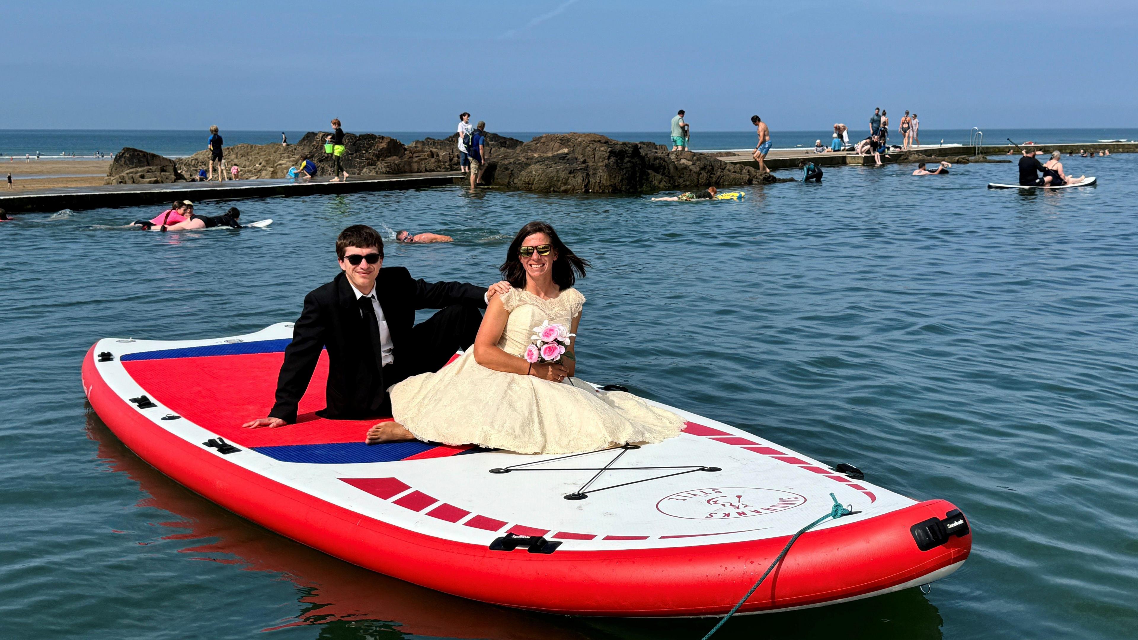 A man in a dark suit and a woman in a cream-coloured dress holding a posy of flowers sit on a paddleboard floating on Bude Sea Pool with swimmers behind them.