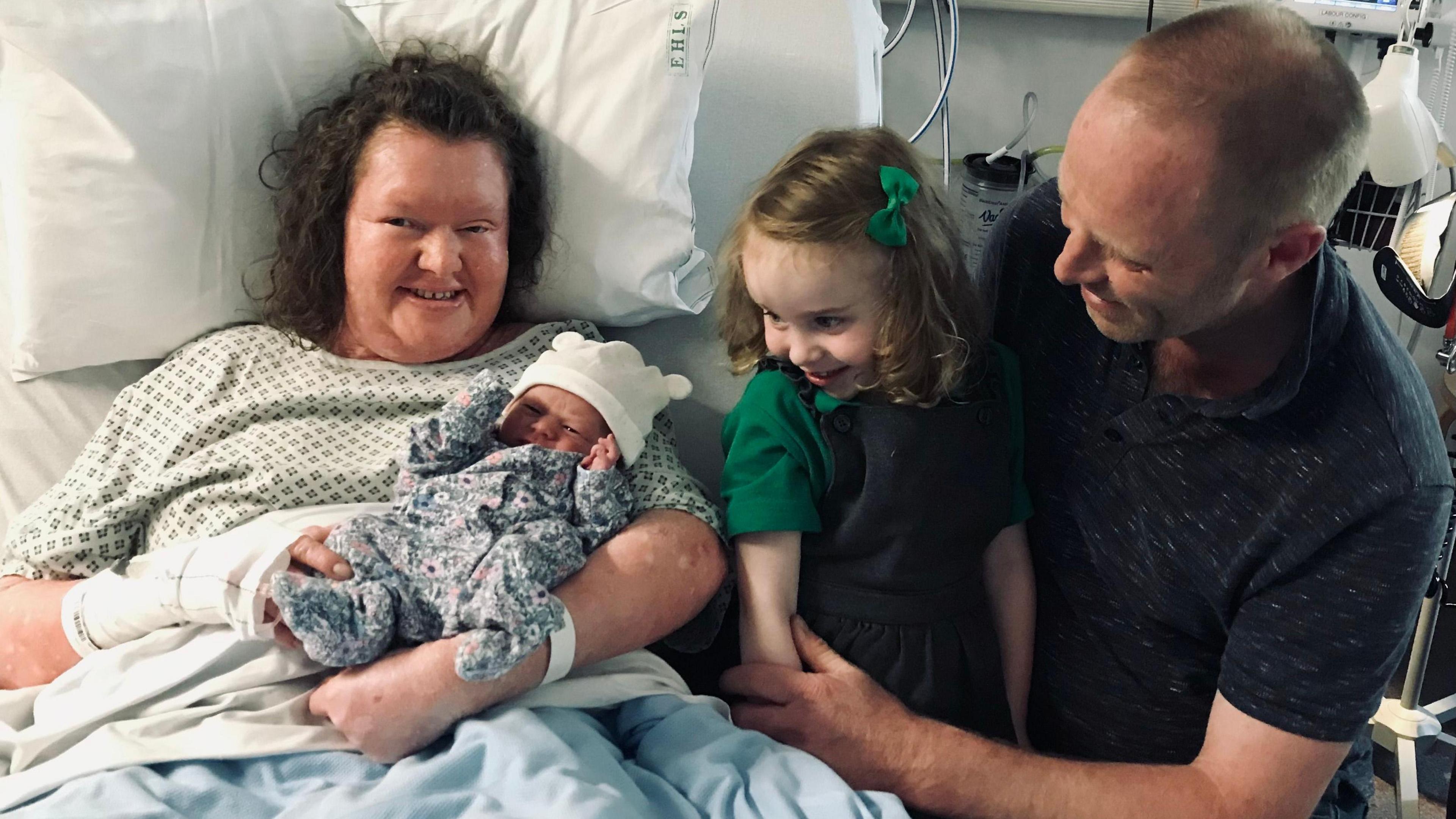 A woman in a hospital bed holding a new born baby with her daughter and husband nearby