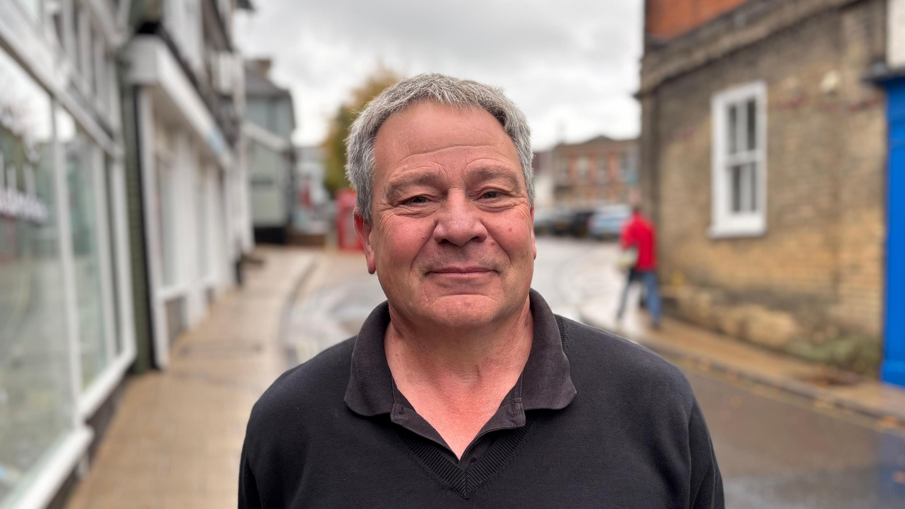 A head and shoulders image of Roger Tripp. He is wearing a black polo shirt and standing on a pavement next to a road. He is looking into the camera and smiling.