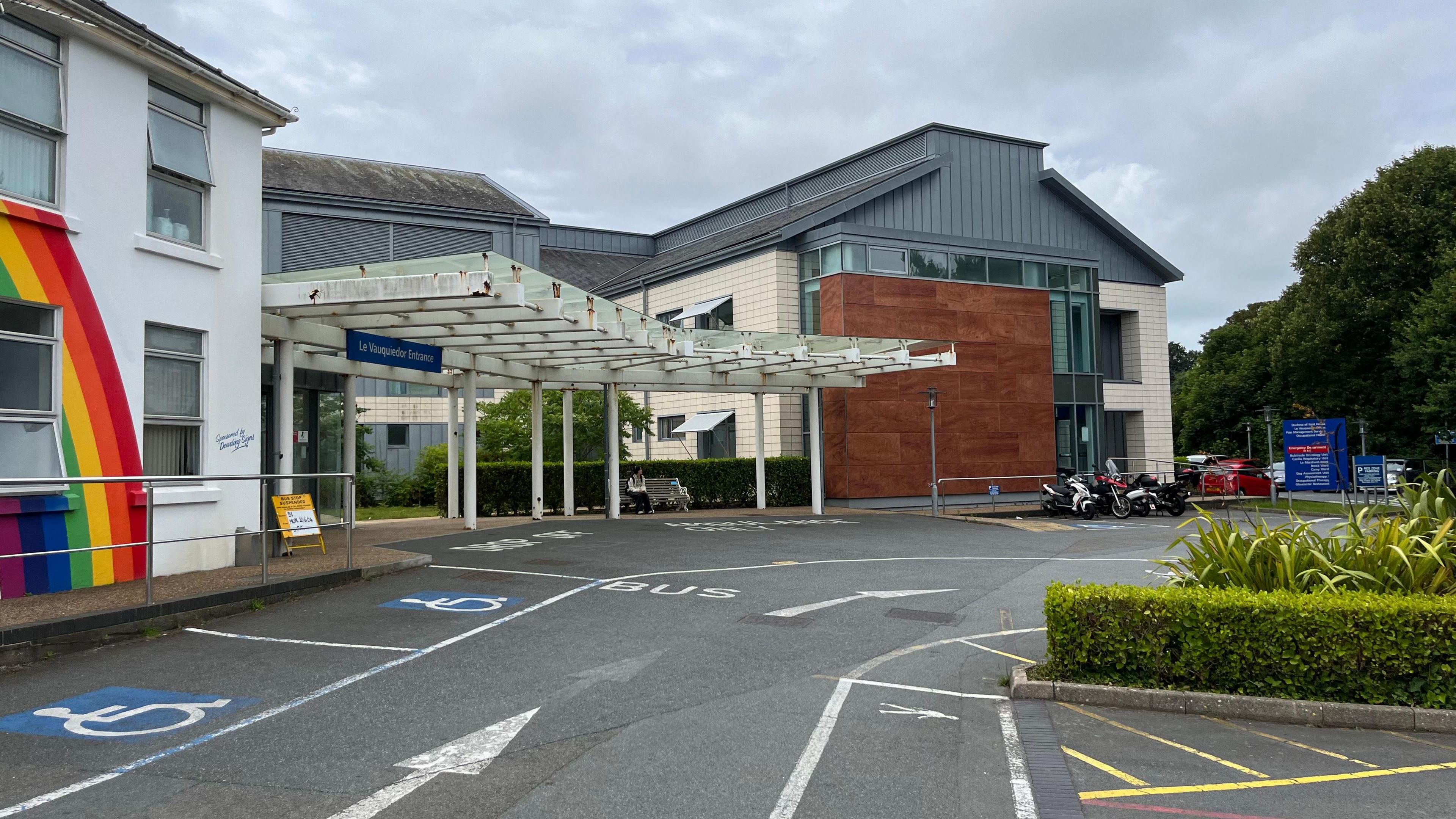 Guernsey's Princess Elizabeth Hospital - A white building with a ranbow painted on it. Next to it is a large criss crossed awning, above a sign which says Le Vauqueidor Entrance. 