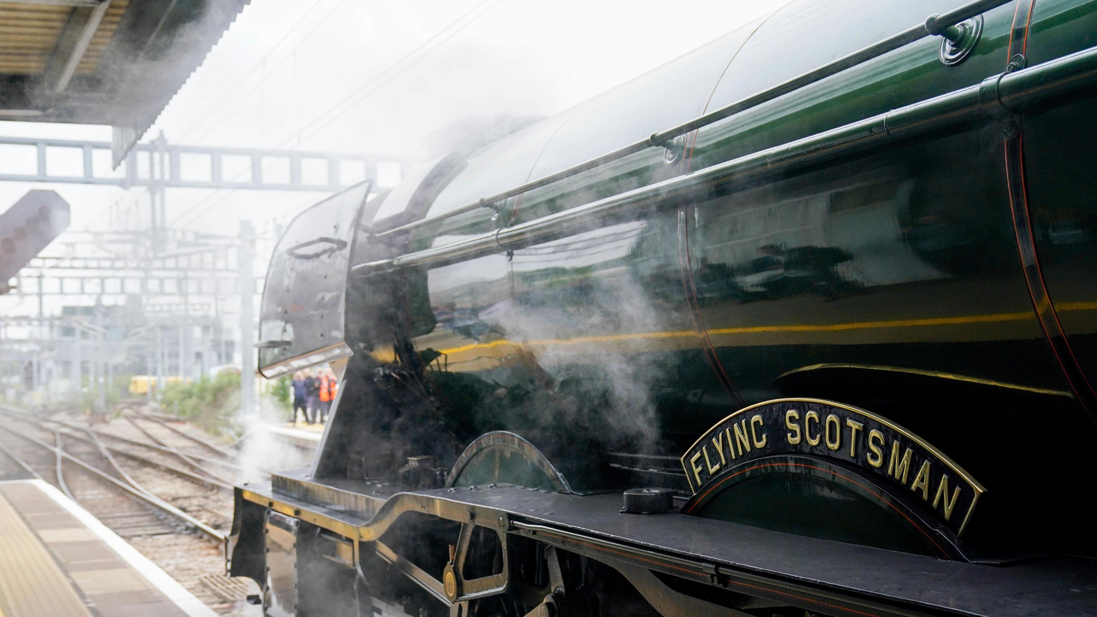 A green and black steam engine works up steam in a station, with a platform to the left and railway lines, with overhead electricity lines, disappearing towards the horizon. The train has a curved named plate which reads, in gold letters on a black background, "Flying Scotsman".