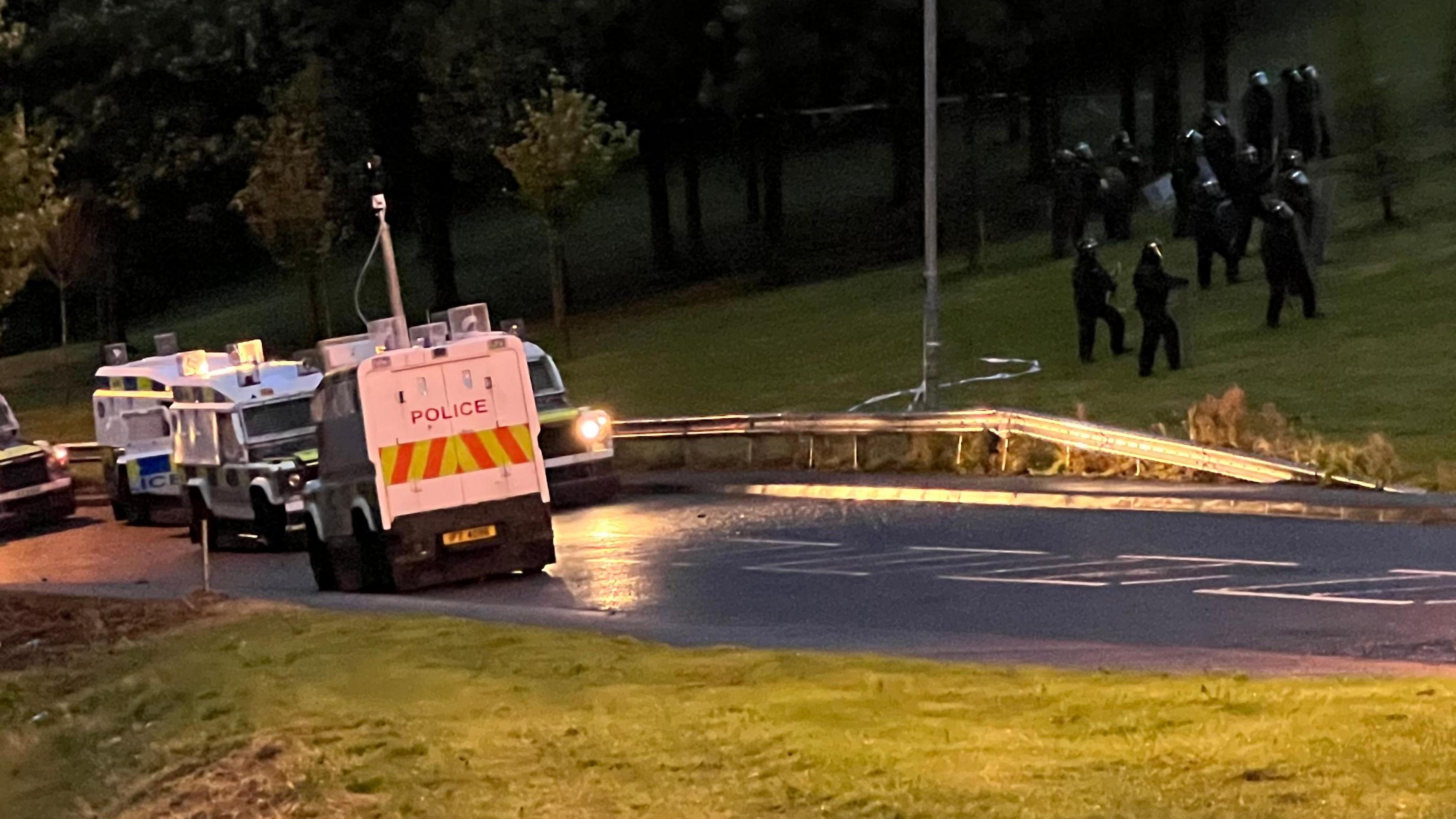 Police landrovers are parked on Southway in Creggan, while officers in protective uniform make their way up a steep banking to the right of the vehicles