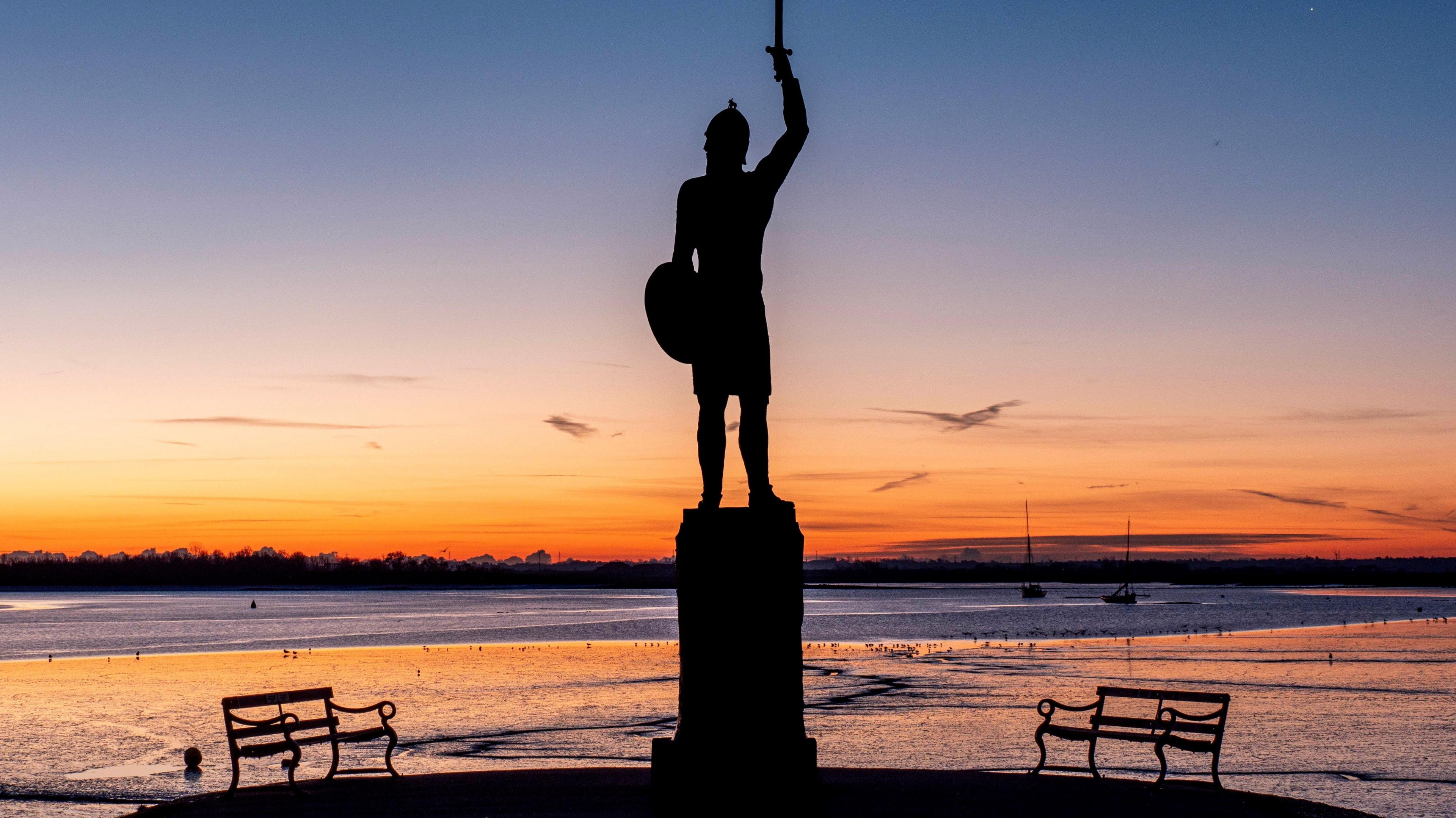 A photo of Byrhtnoth statue in Maldon's Promenade Park, showing a warrior holding up a sword on his right arm and a shield in his left. It is in silhouette with a bench either side, a golden horizon and the Blackwater estuary in front, with two ships in the distance. 