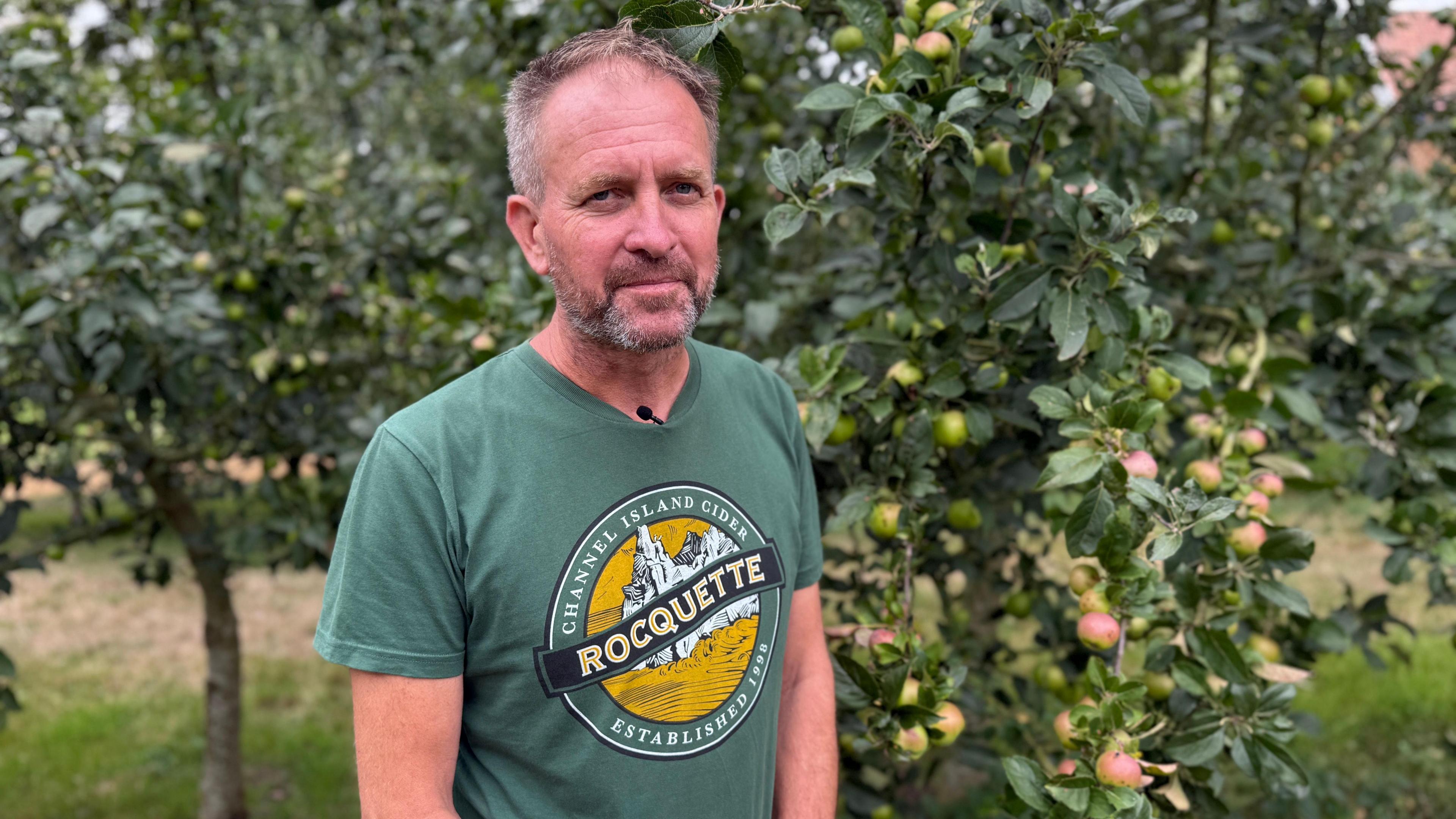 James Meller with short hair looking at the camera. He is wearing a green t-shirt with the logo for Rocquette Cider on the front. A tree with small apples is in the background