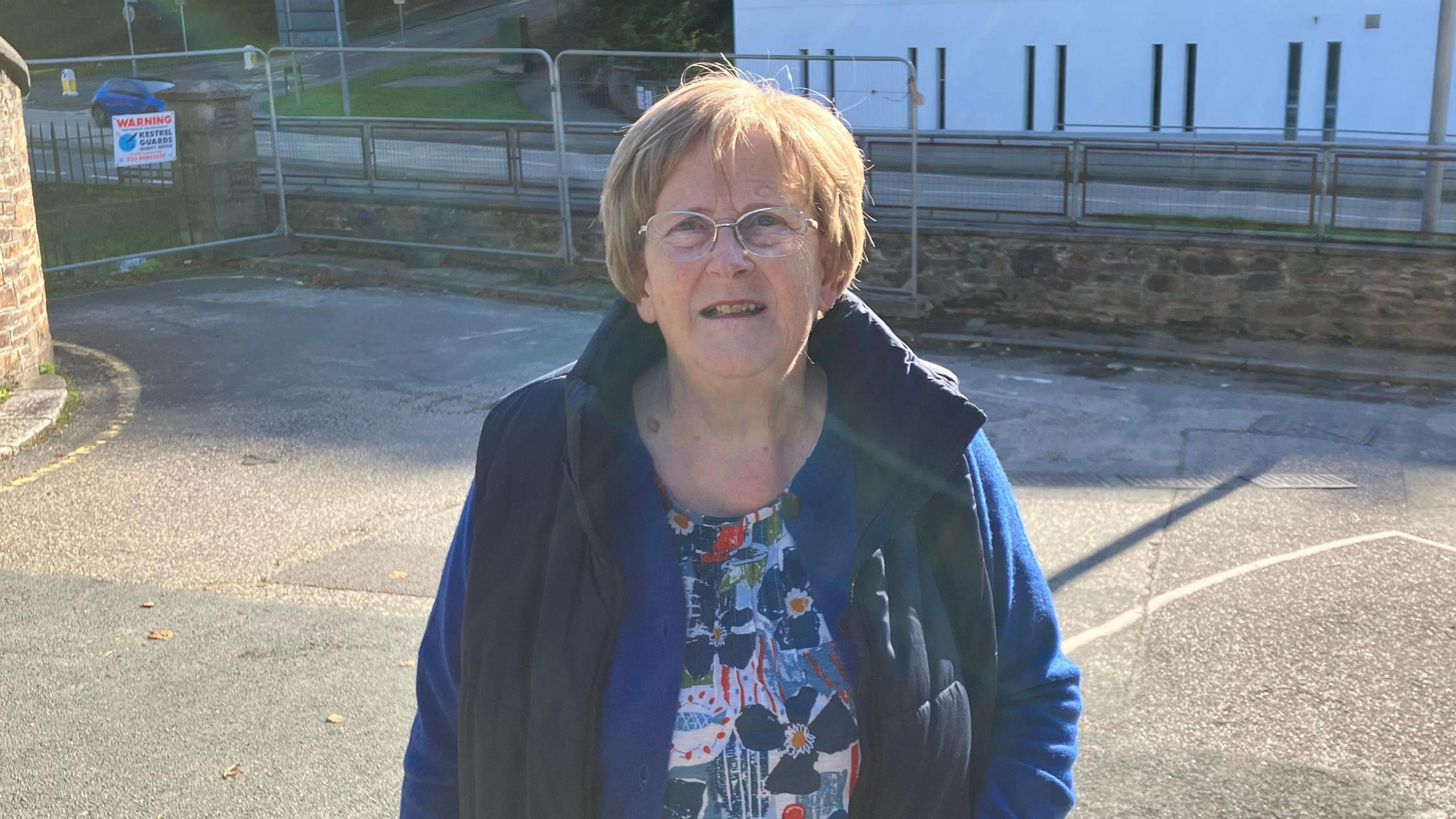A woman with red hair and glasses wearing a dark blue body warmer, purple cardigan, and flowery top with Tregolls Road and a metal wire fence in the background. 
