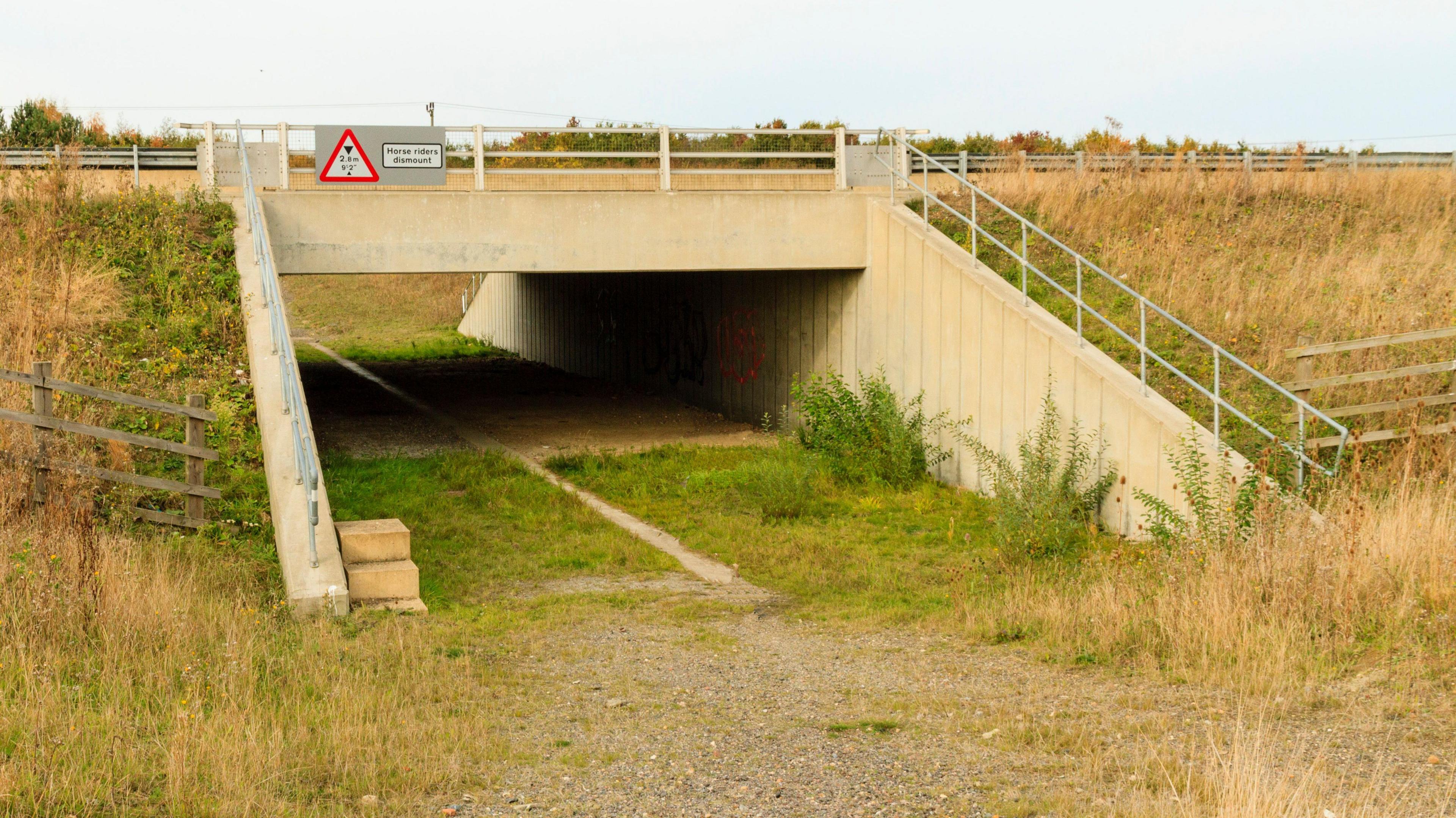 A stone bridge on the A421 in Bedfordshire with railings around it and grasses on the banks either side. There is a path with greenery underneath it. 